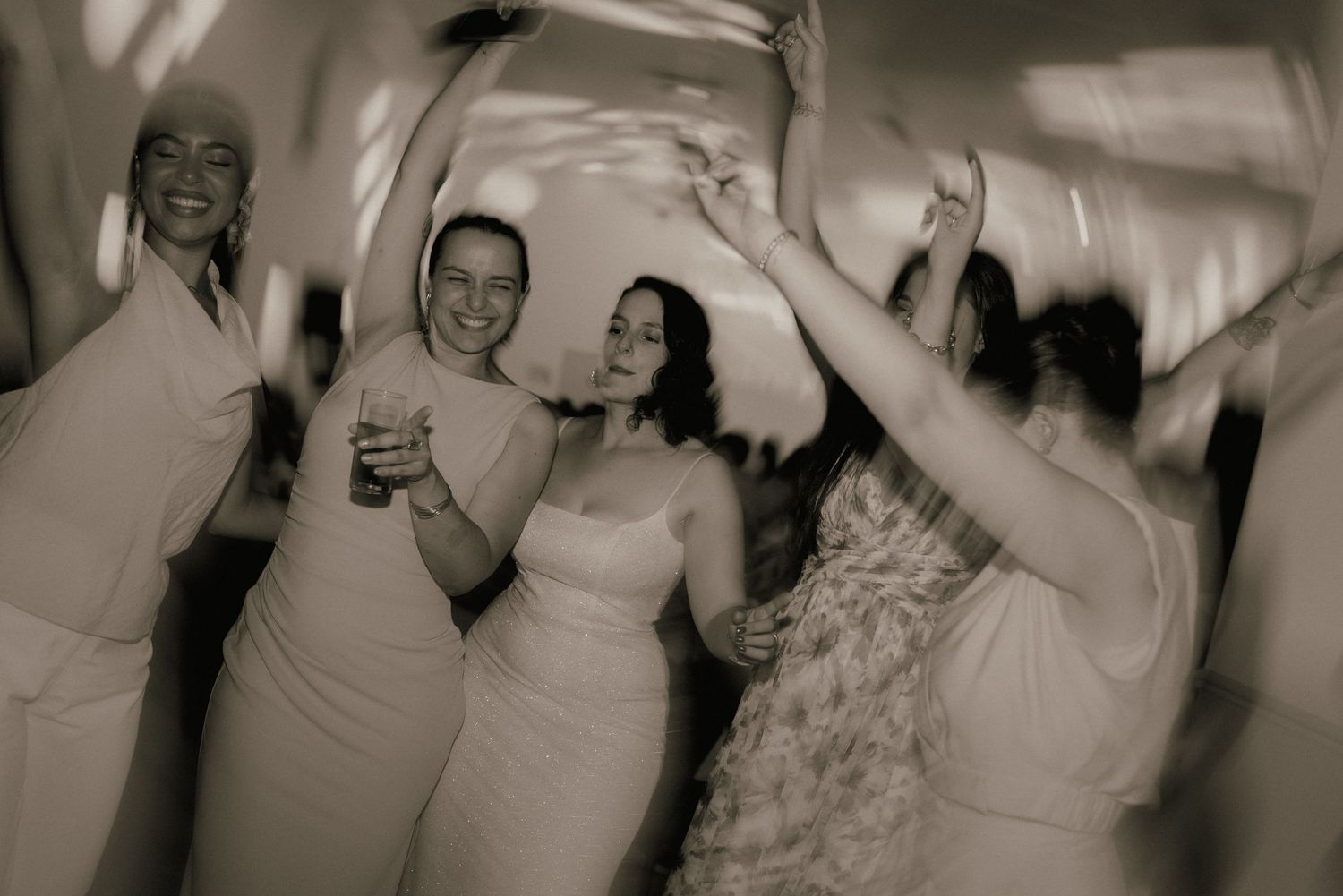 Black and white photo of bride and bridesmaids dancing joyfully at wedding reception.
