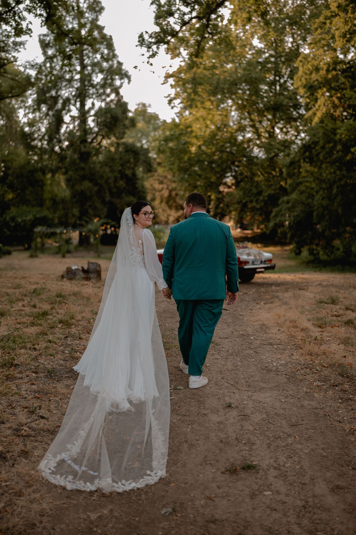Bride in white gown with long veil walks hand-in-hand with groom in teal suit along a tree-lined dirt path.