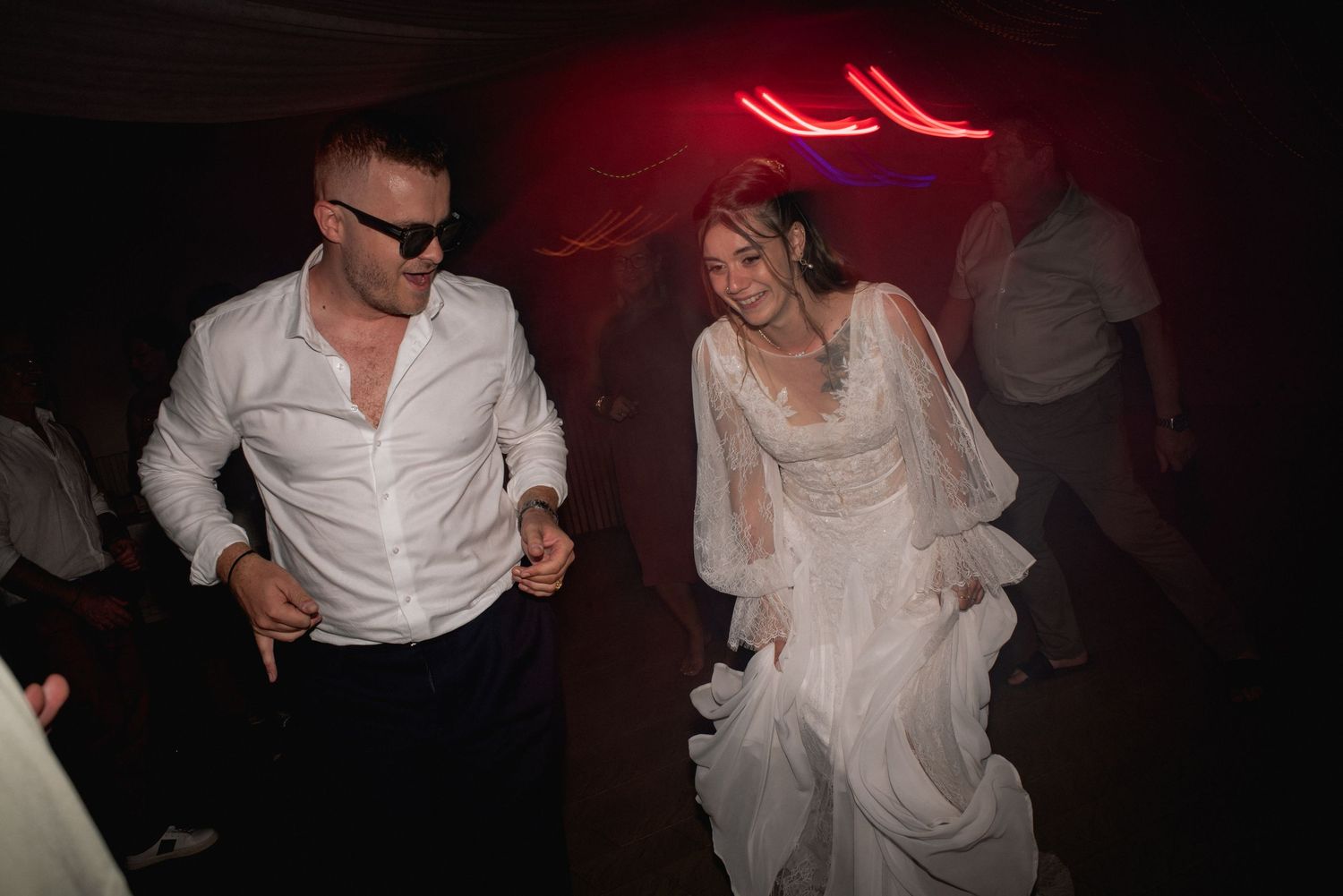Bride in white lace gown and groom in white shirt dancing joyfully at a wedding reception.