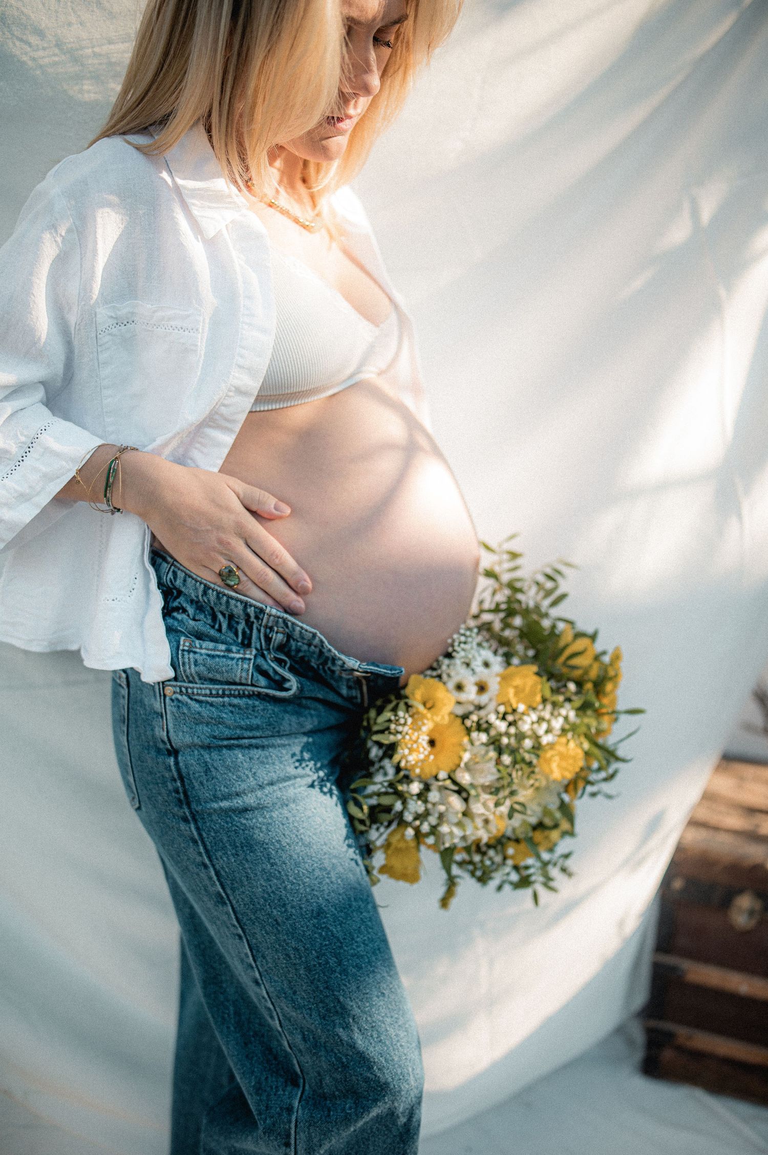 Pregnant woman in white top and jeans holding yellow wildflowers, cradling bare baby bump.