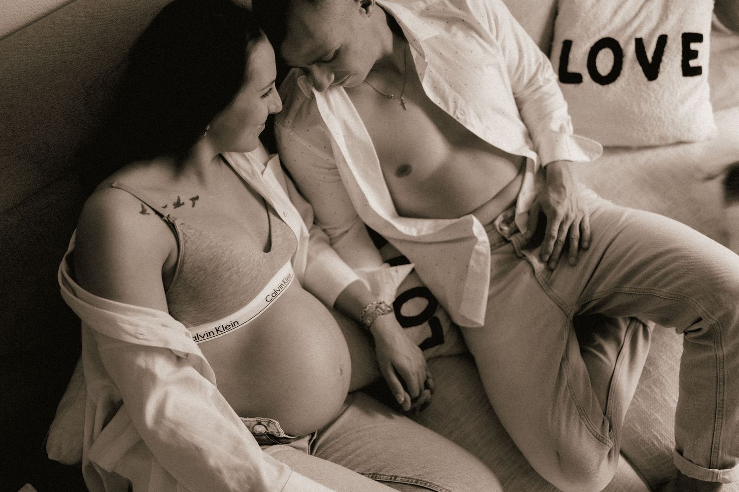 Intimate sepia maternity photo of couple reclining together with pregnant belly and LOVE pillow.