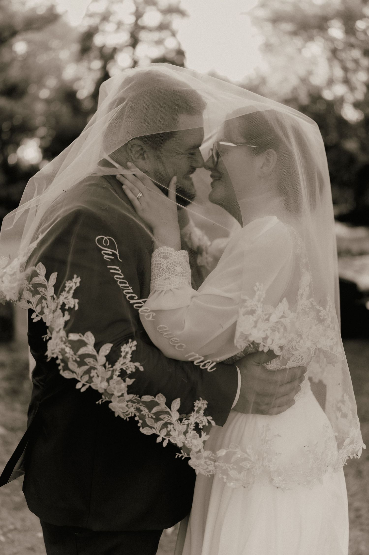 Black and white photo of bride and groom sharing intimate moment under lace veil with floral trim.