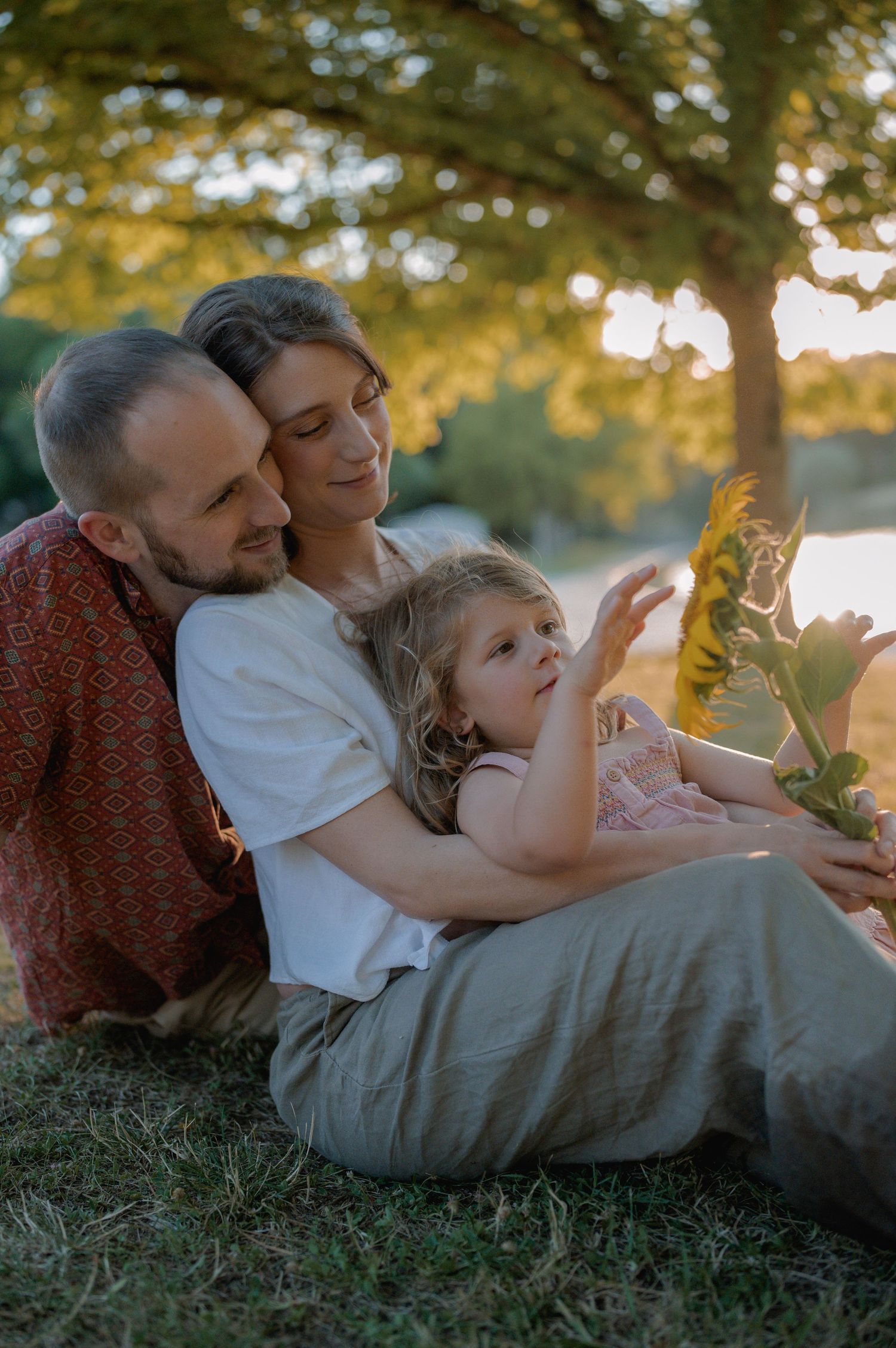 A family of three sits together in golden hour light under a tree, watching their toddler explore.