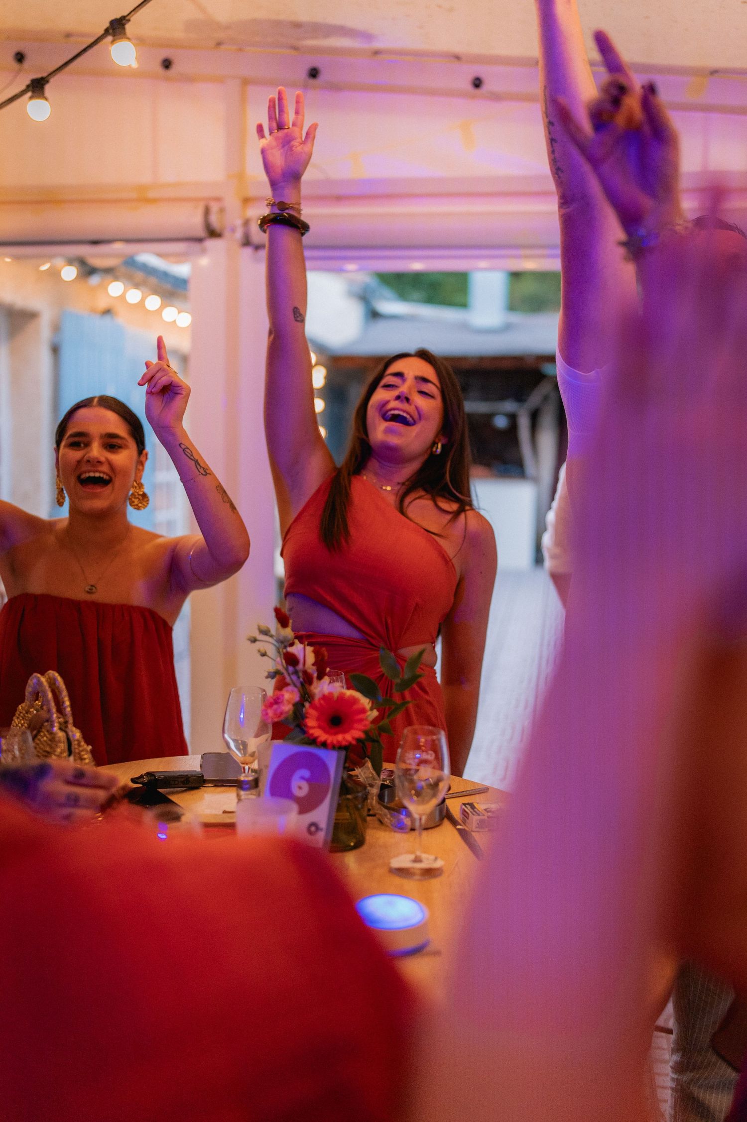 Women in red dresses celebrate at a wedding reception, dancing with hands raised under warm string lights.