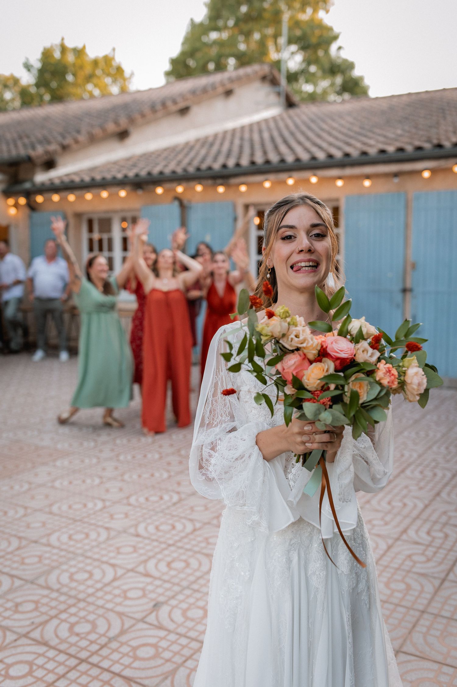 Smiling bride in white dress holds colorful bouquet with cheering wedding guests celebrating in background.