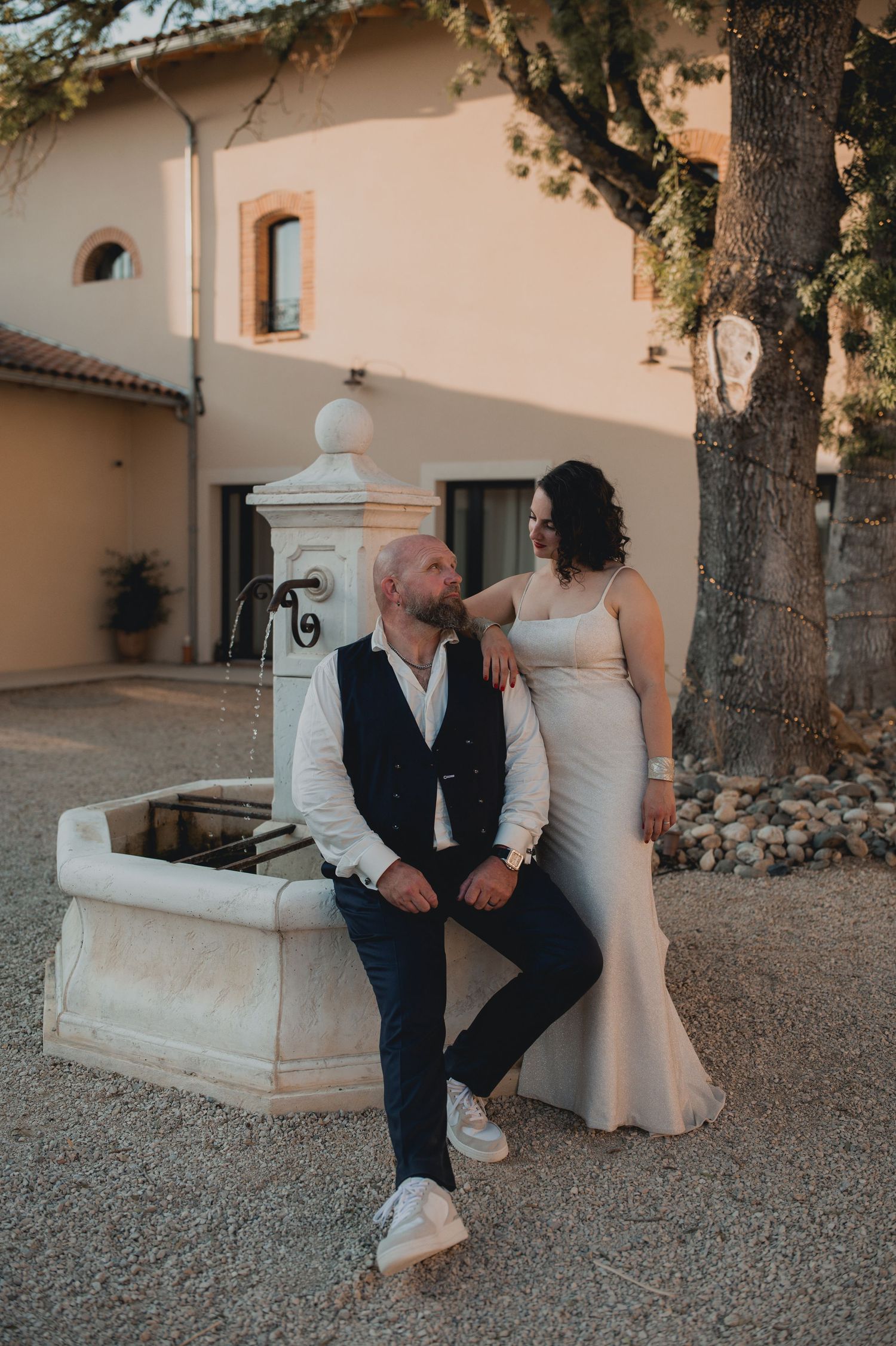 A couple in wedding attire poses by a stone fountain near a rustic villa, sharing a romantic moment at golden hour.
