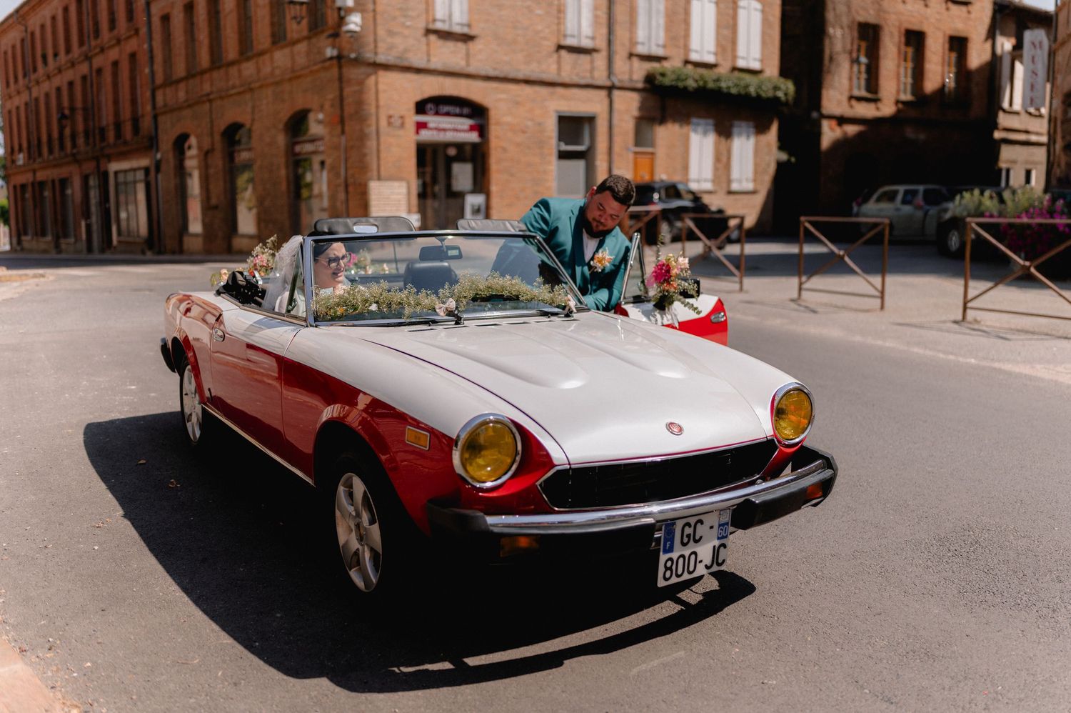 A classic red and white Fiat 124 Spider convertible parked on a sunny street with a person leaning into the car.