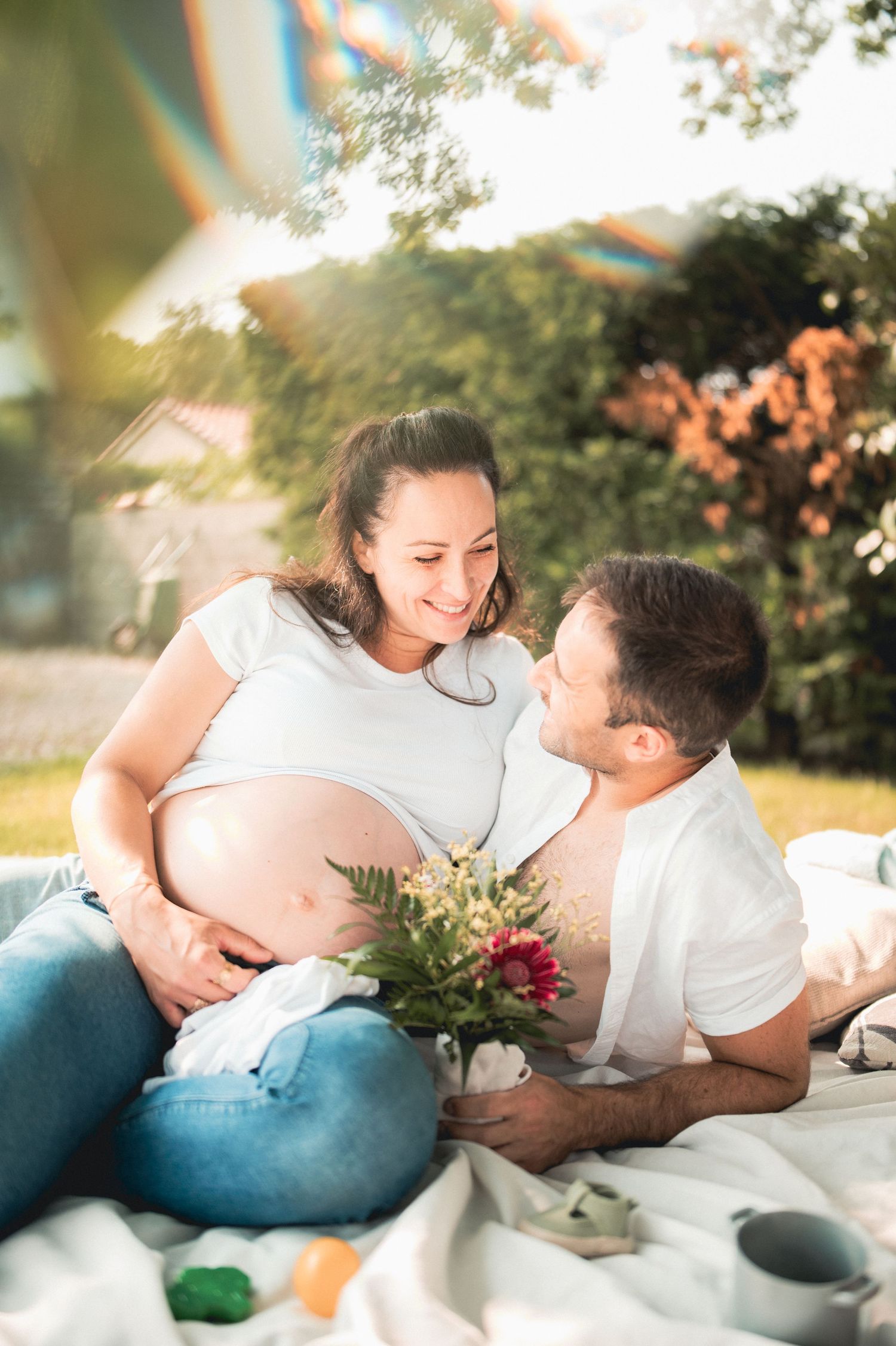 Pregnant couple relaxing on blanket in sunny garden, smiling at each other with wildflower bouquet.