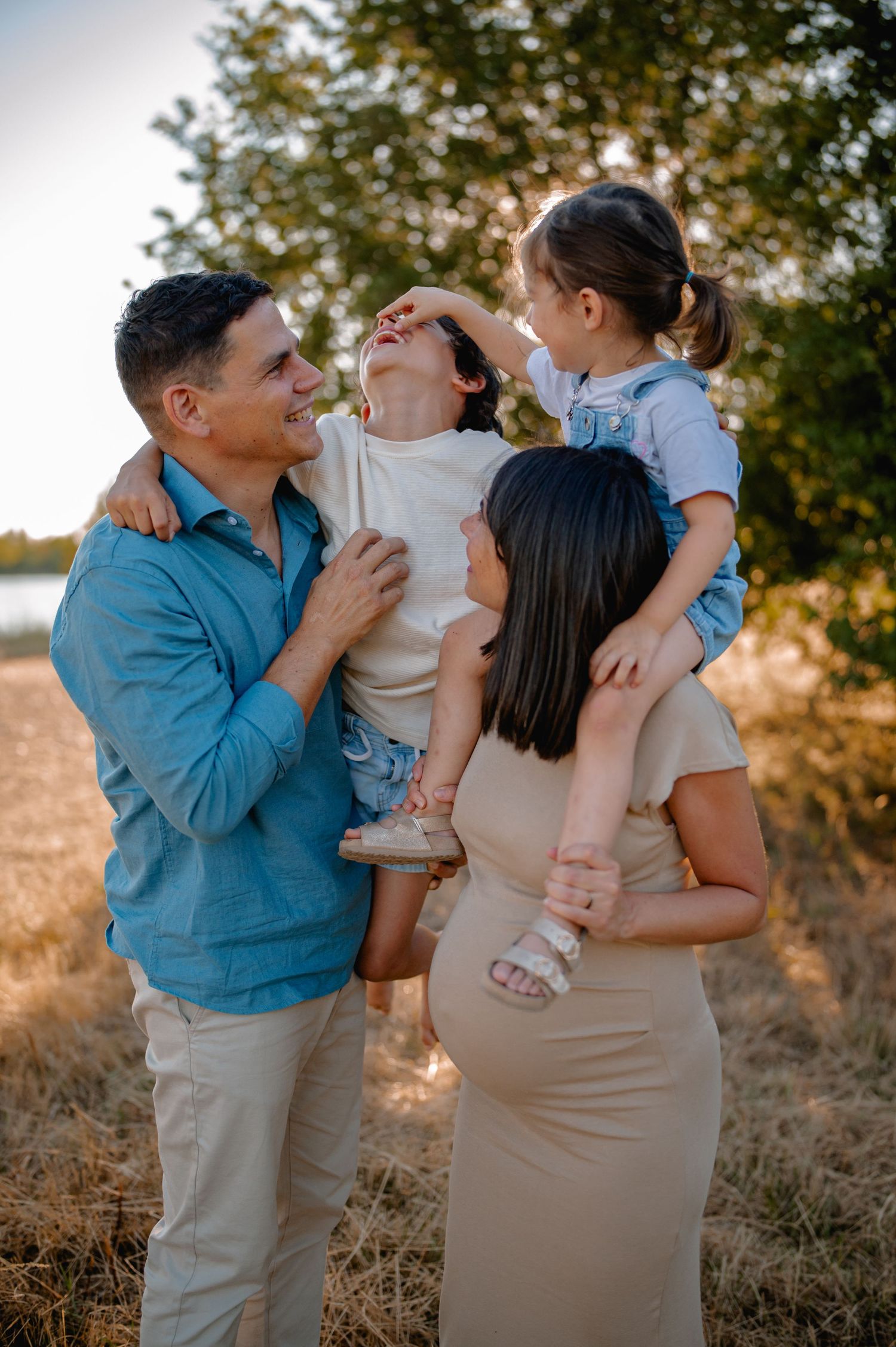 Joyful pregnant family of four sharing a playful moment together in a sun-drenched field.