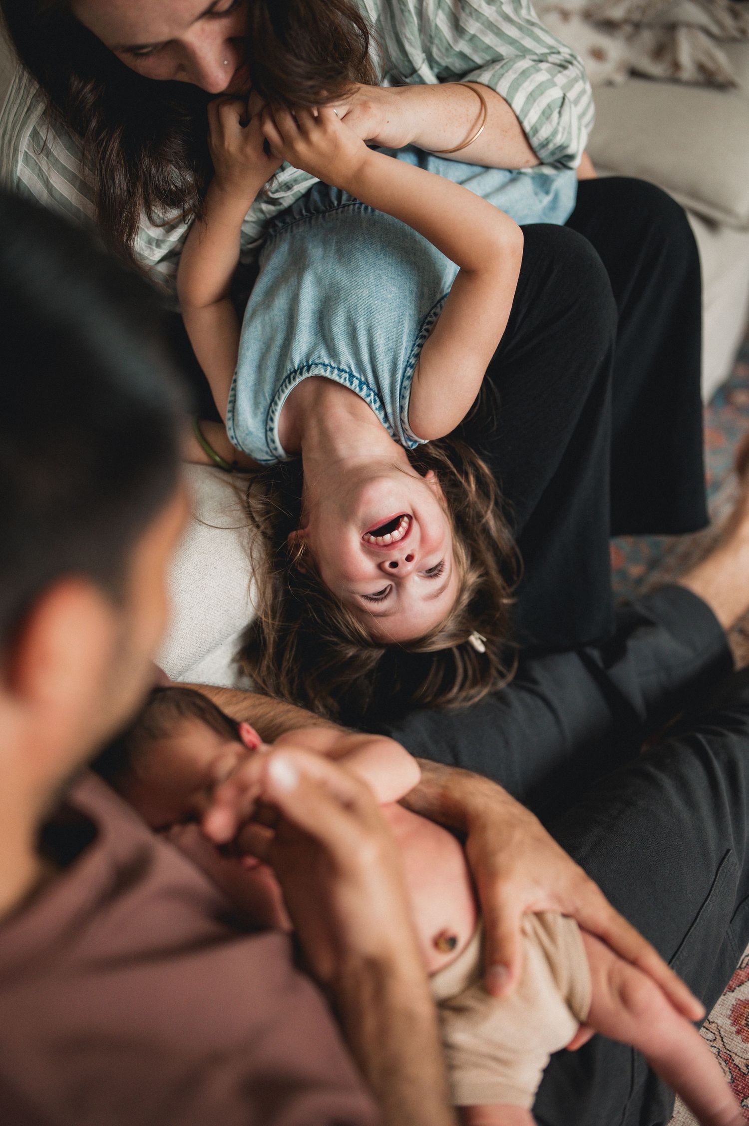 Laughing child lying upside down on mothers lap while father holds newborn in candid family photo.