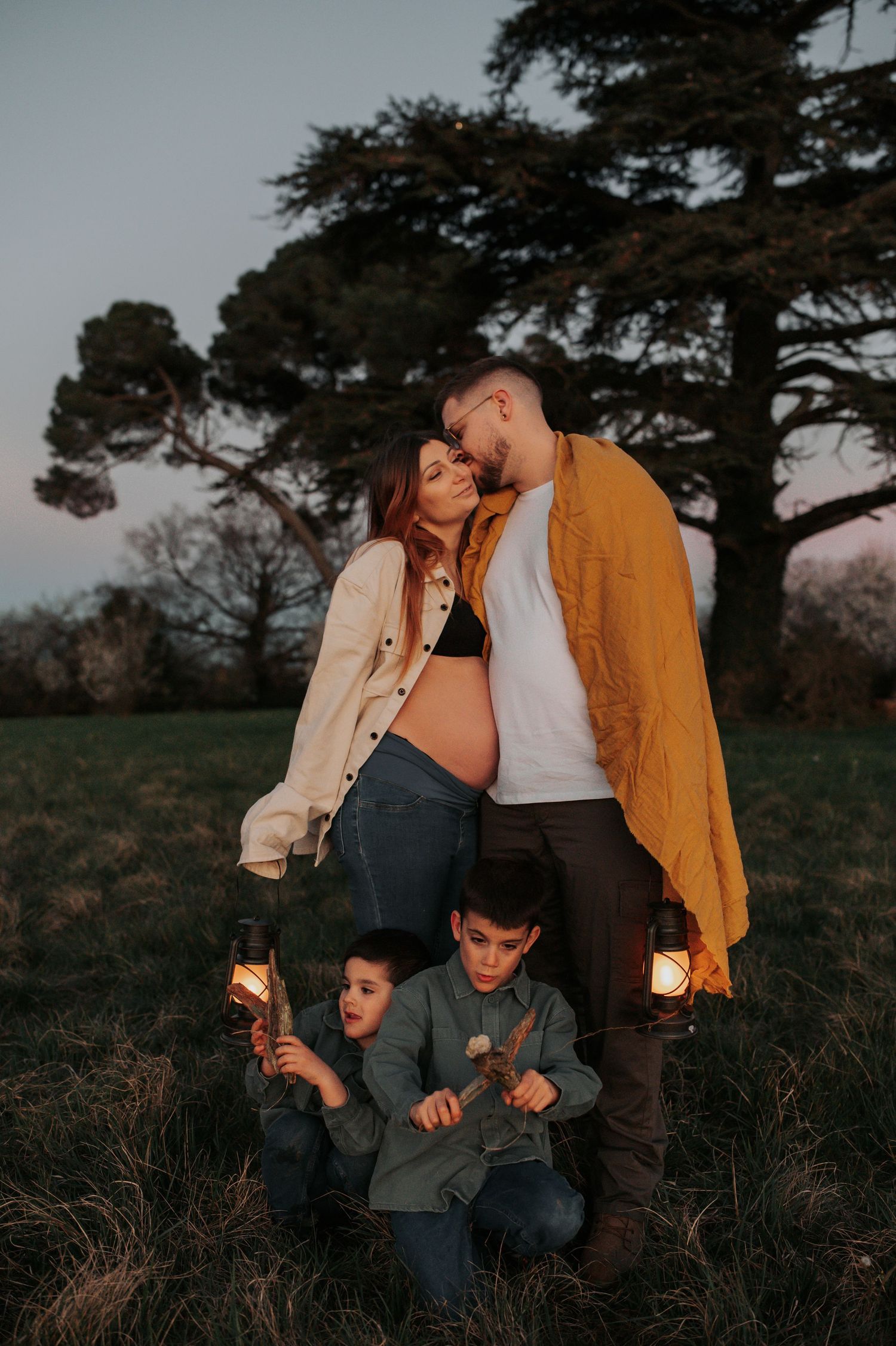 Family maternity photo at dusk with pregnant mother, father, and two children holding lanterns in a field.