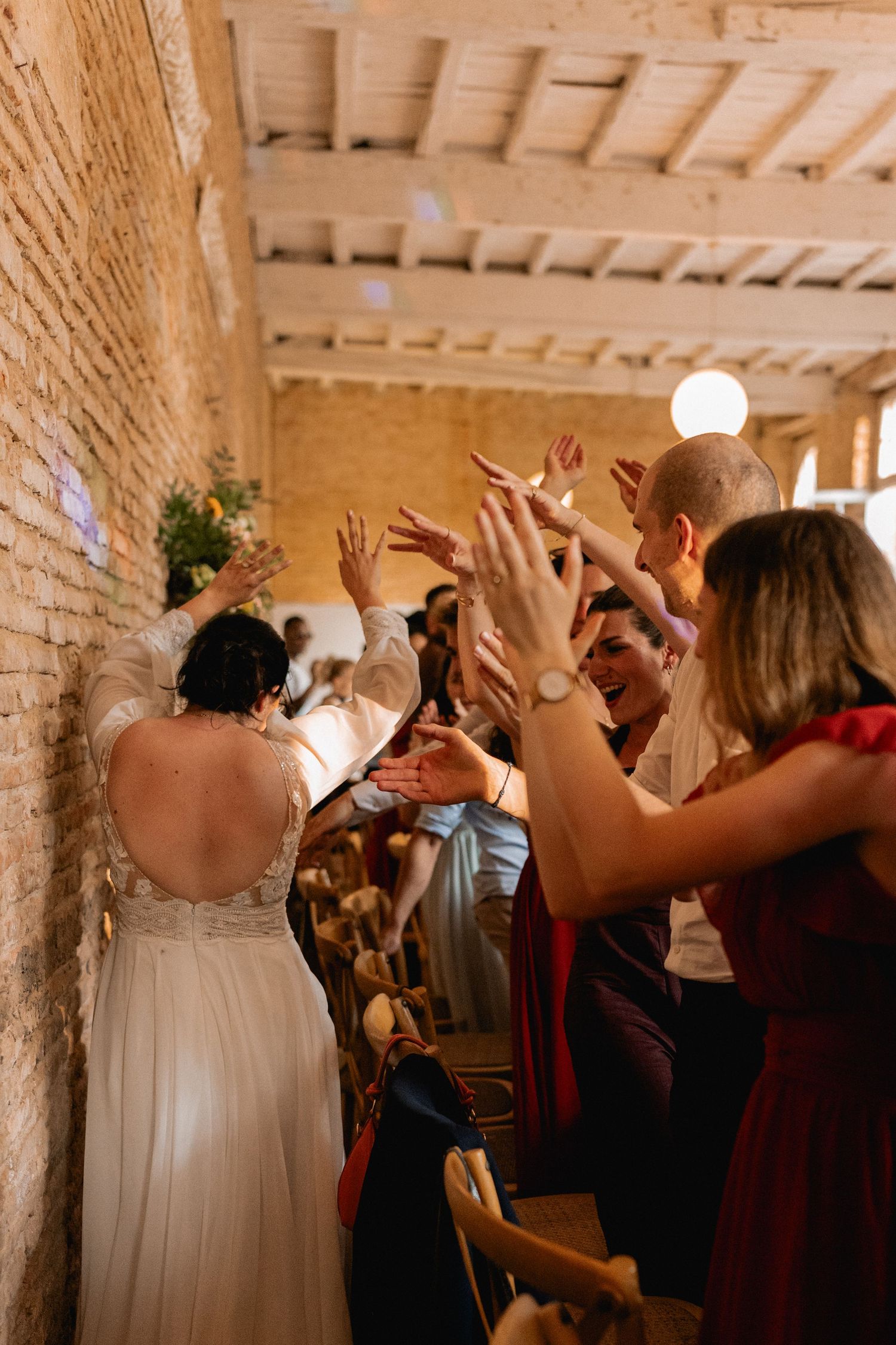 Bride in white gown walks through cheering guests with raised hands at a rustic wedding venue.