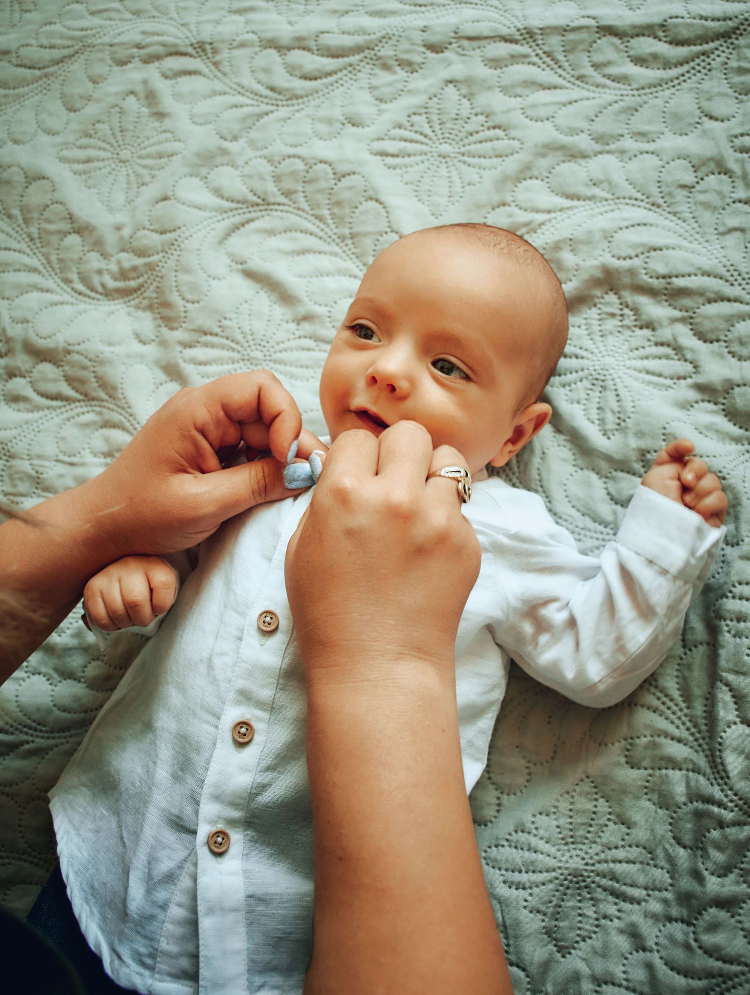 Newborn baby in white shirt lying on quilted blanket while parent buttons up outfit with ringed hands.