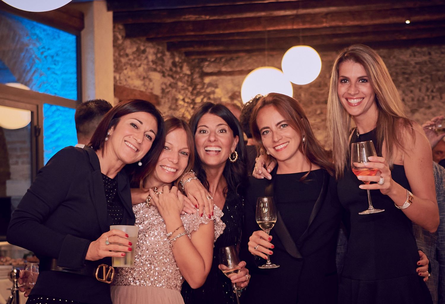 Five women in elegant attire smile and hold champagne glasses at a festive indoor celebration.