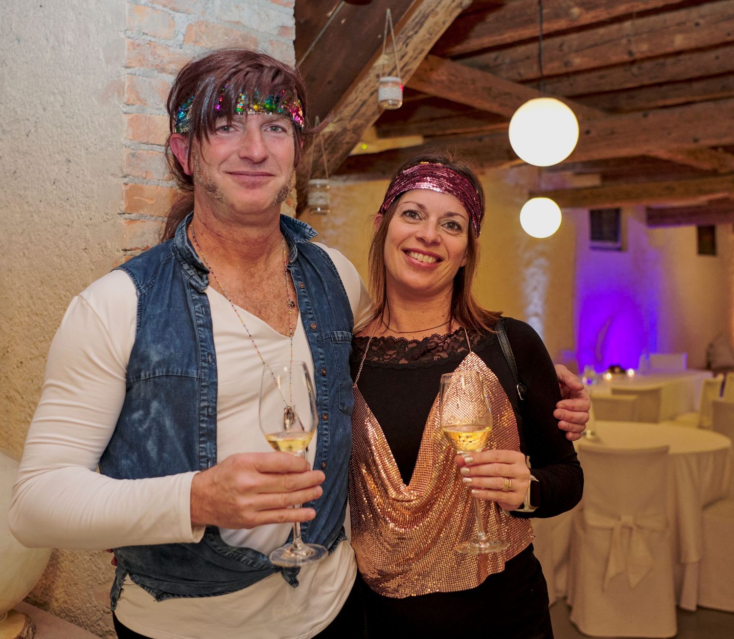 A man and woman in festive costumes hold champagne glasses at a themed indoor celebration.