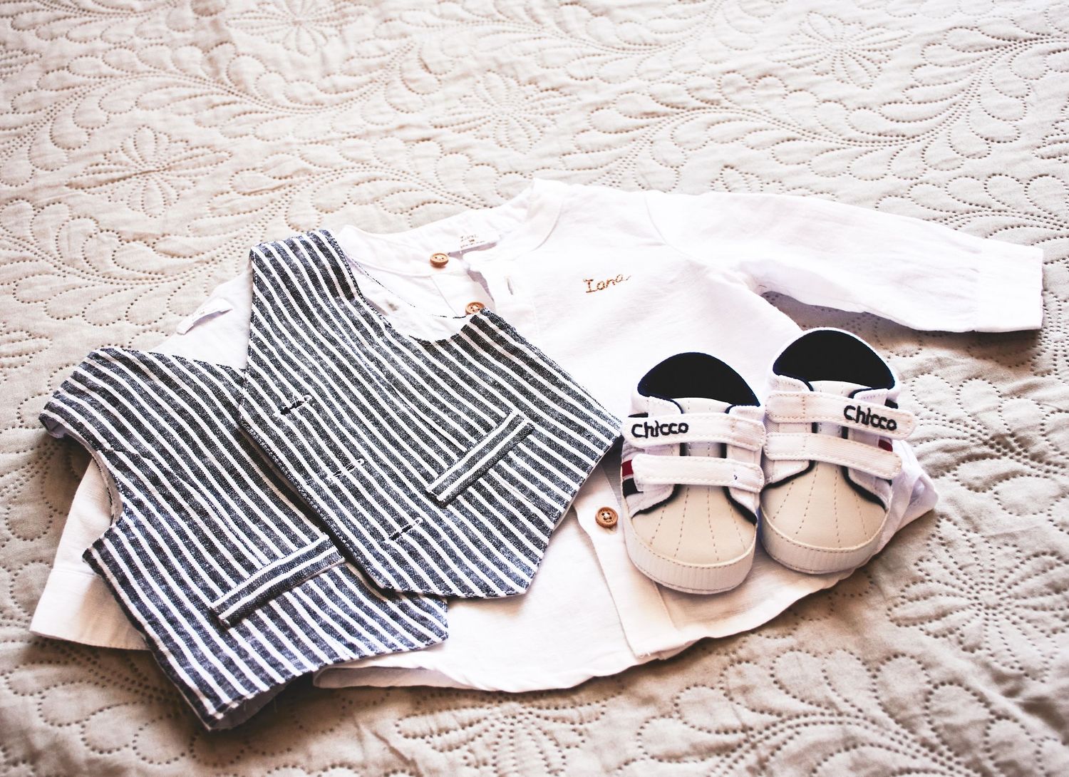 Baby boy christening outfit with striped vest, white shirt, and Chloé sneakers laid flat on a quilted white surface.