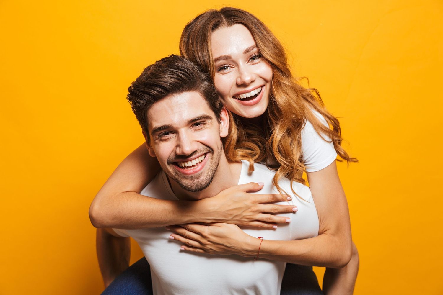 A happy couple smiling as the woman gives the man a piggyback ride on a yellow background.