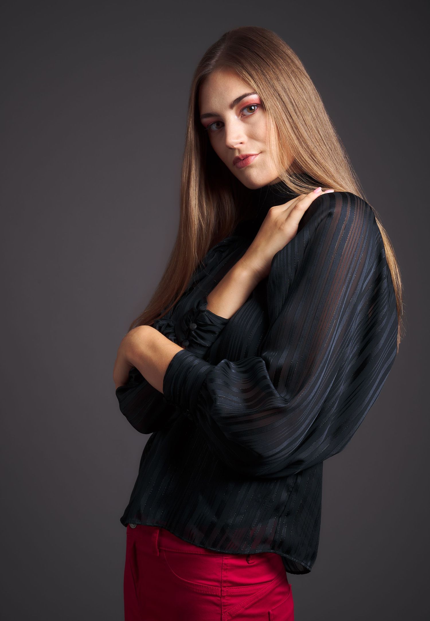 Young woman with long brown hair posing in black sheer blouse and red pants against dark background.
