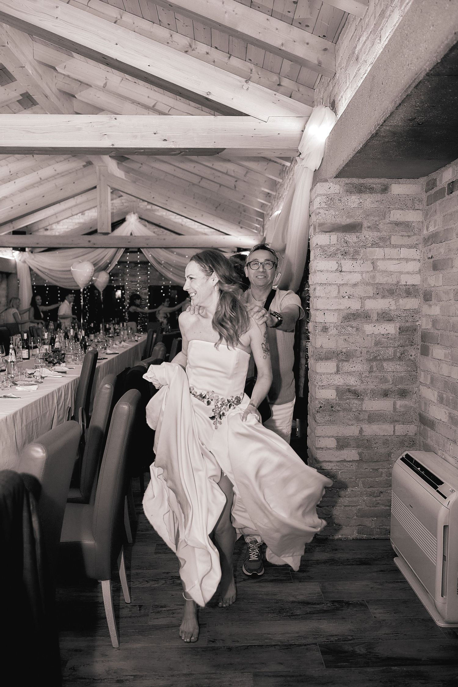 Bride laughs joyfully while being carried at indoor wedding reception.