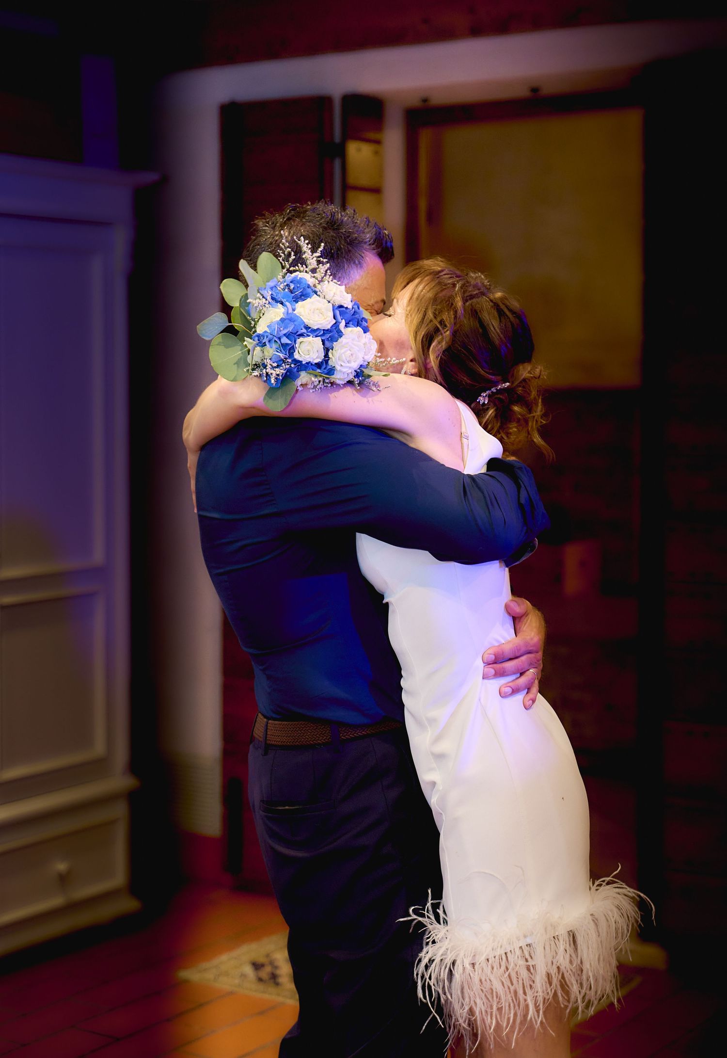 Bride and groom share emotional embrace holding blue wedding bouquet.