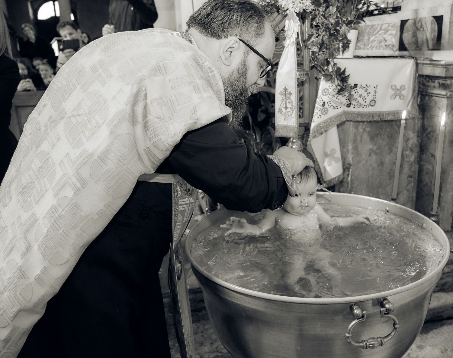 Priest immersing infant in baptismal font during Orthodox christening ceremony in black and white.