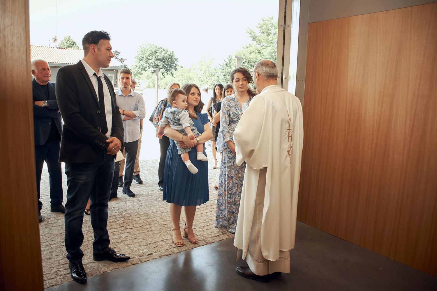 Family greeting a priest at church entrance during a baptism ceremony outdoors.