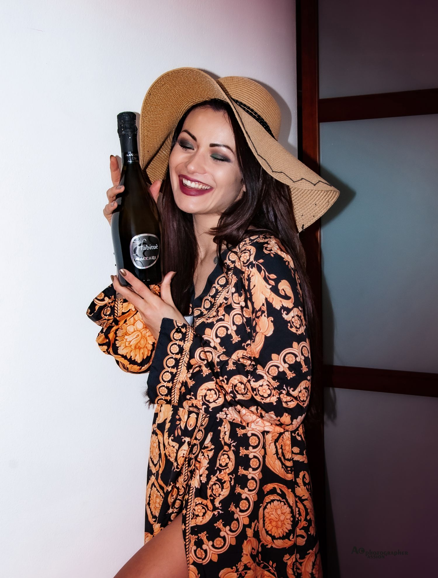 Smiling brunette woman in Versace-print robe and sun hat holding champagne bottle indoors.