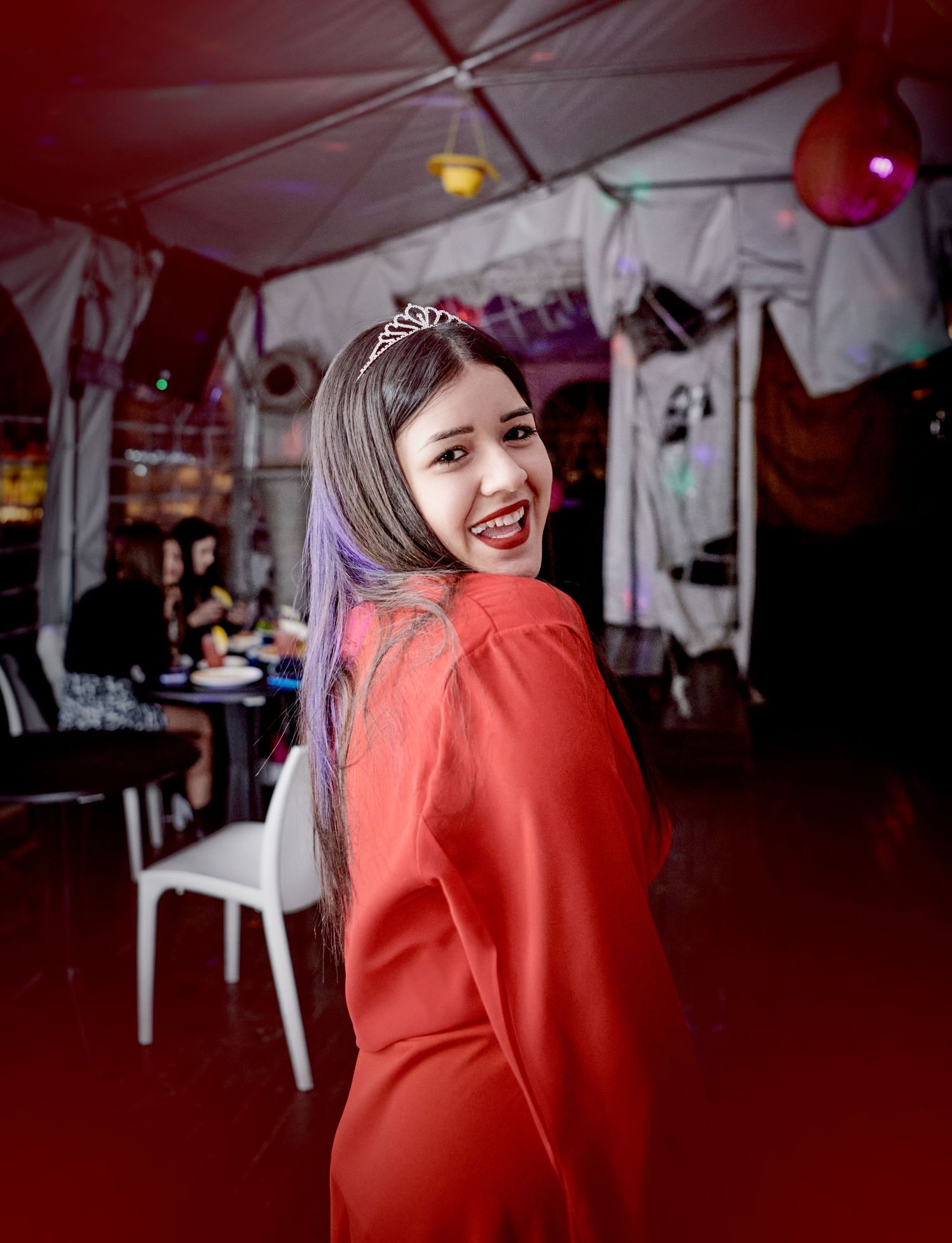 Smiling young woman in red dress wearing tiara at a festive indoor birthday celebration.