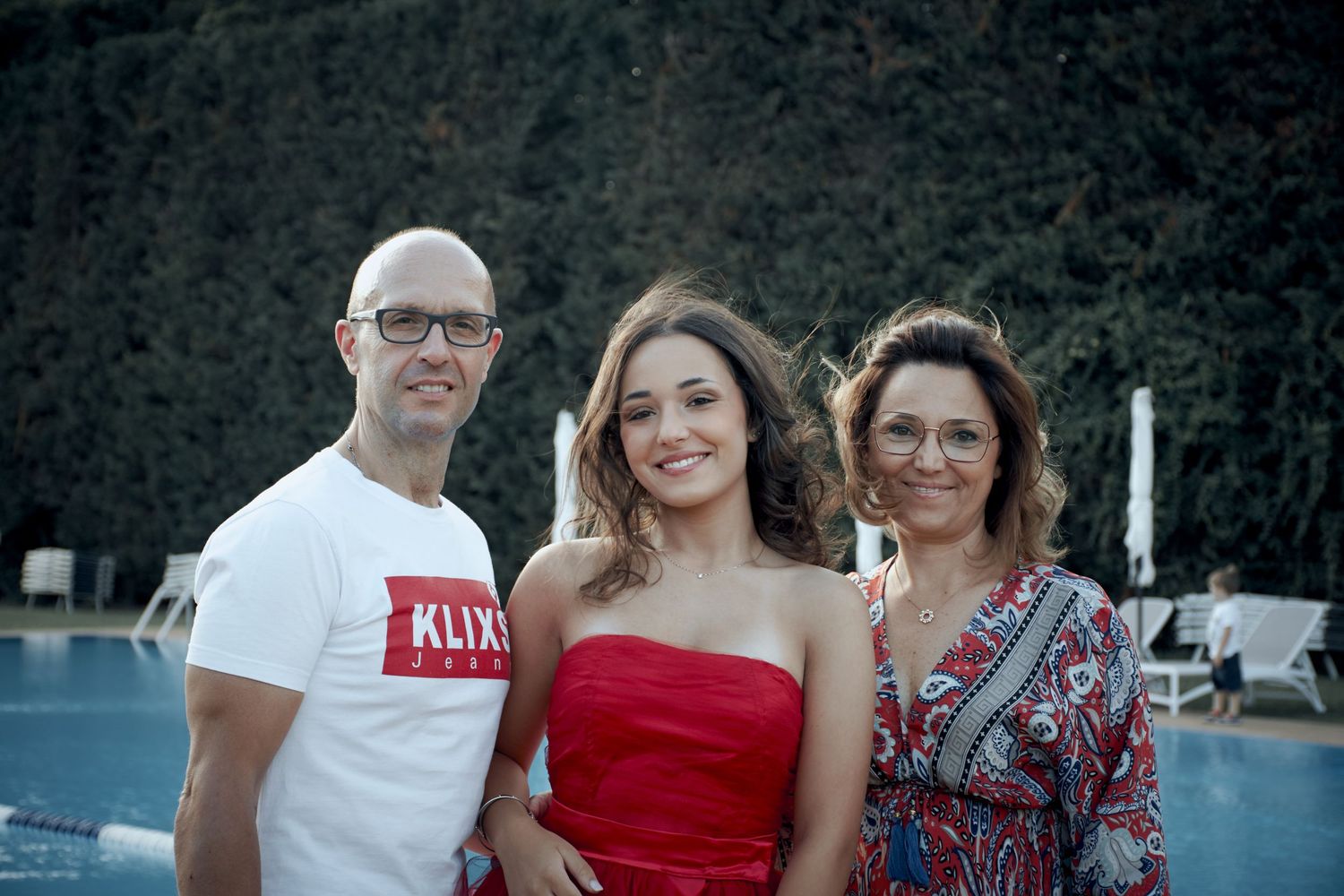 Man in white shirt and woman in floral dress pose with young woman in red dress outdoors.