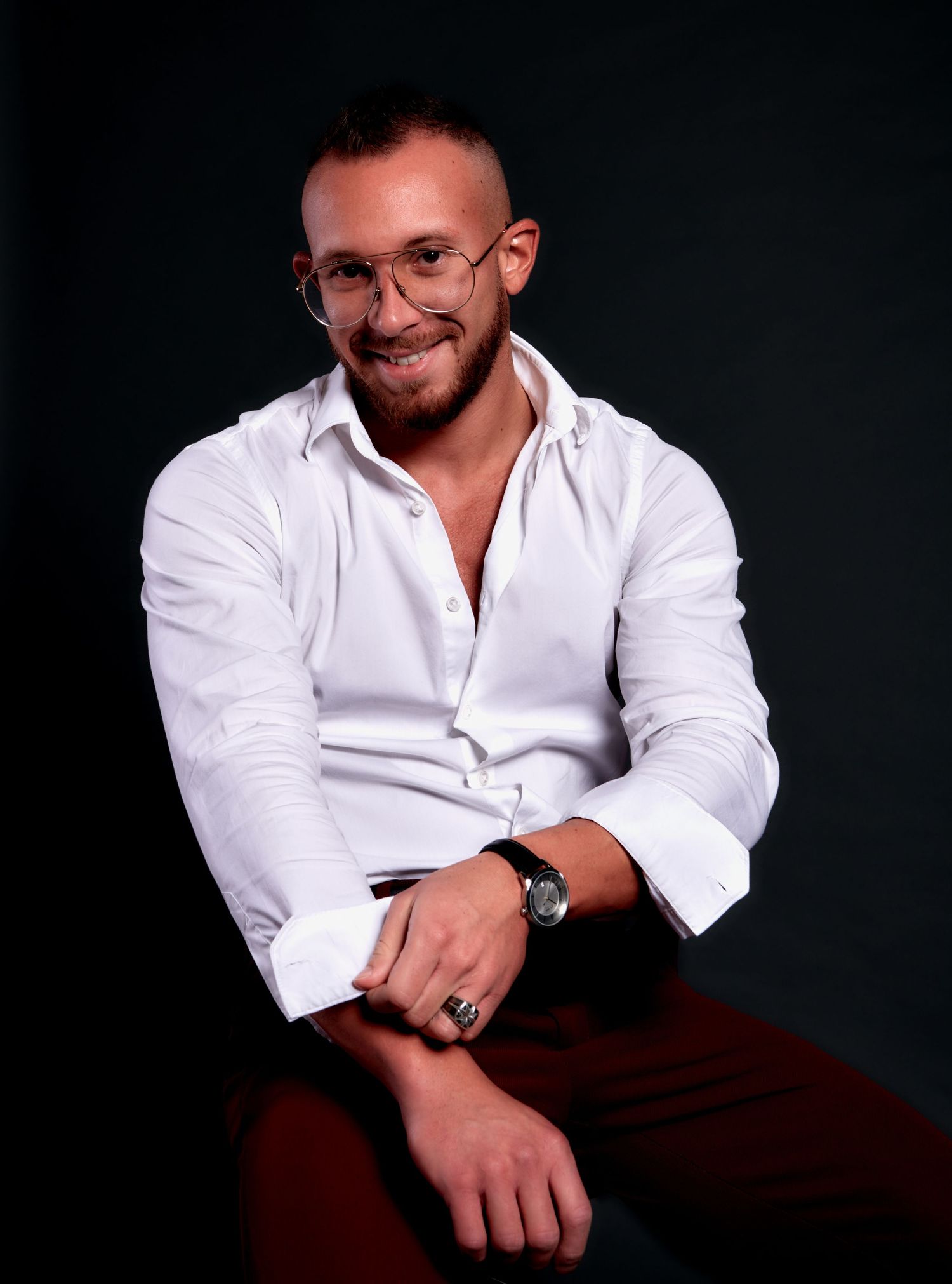 A smiling muscular man in a white shirt and glasses poses confidently against a dark background.