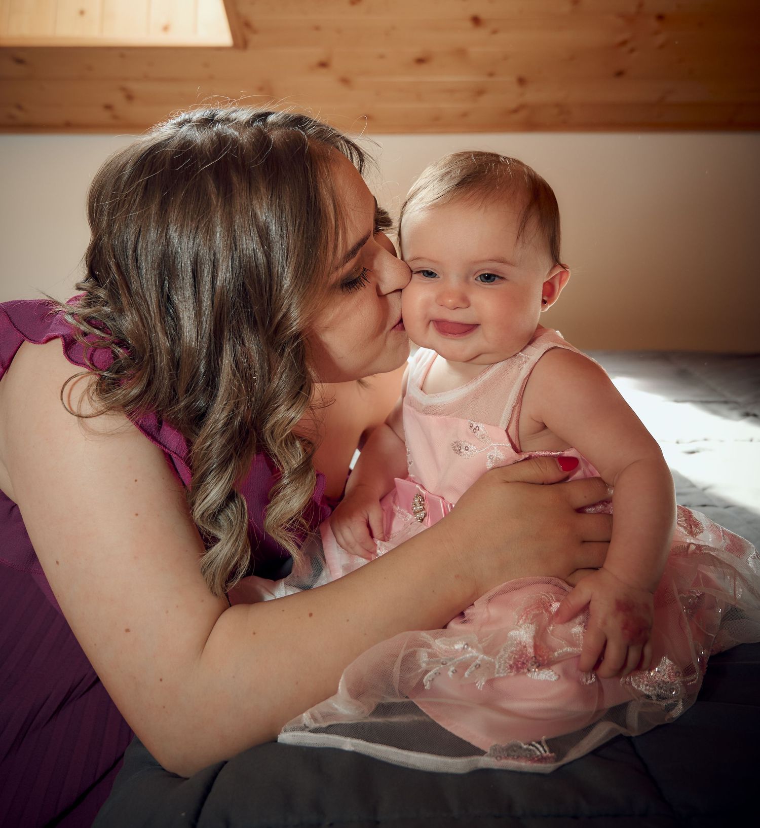 Loving mother with curly hair kissing her smiling baby girl in pink tutu dress on a bed.