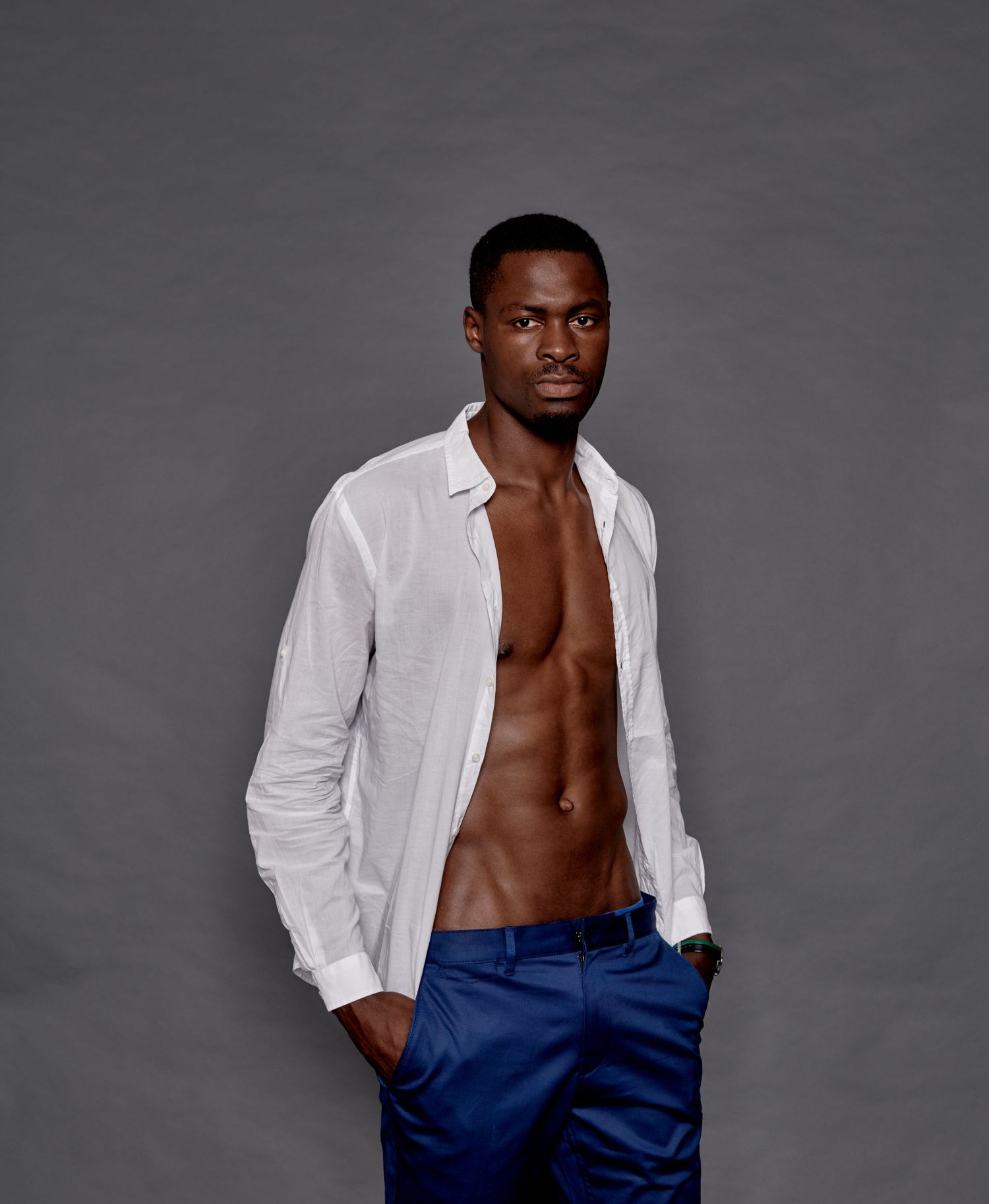 Athletic Black male model in open white shirt and blue pants posing on gray background.