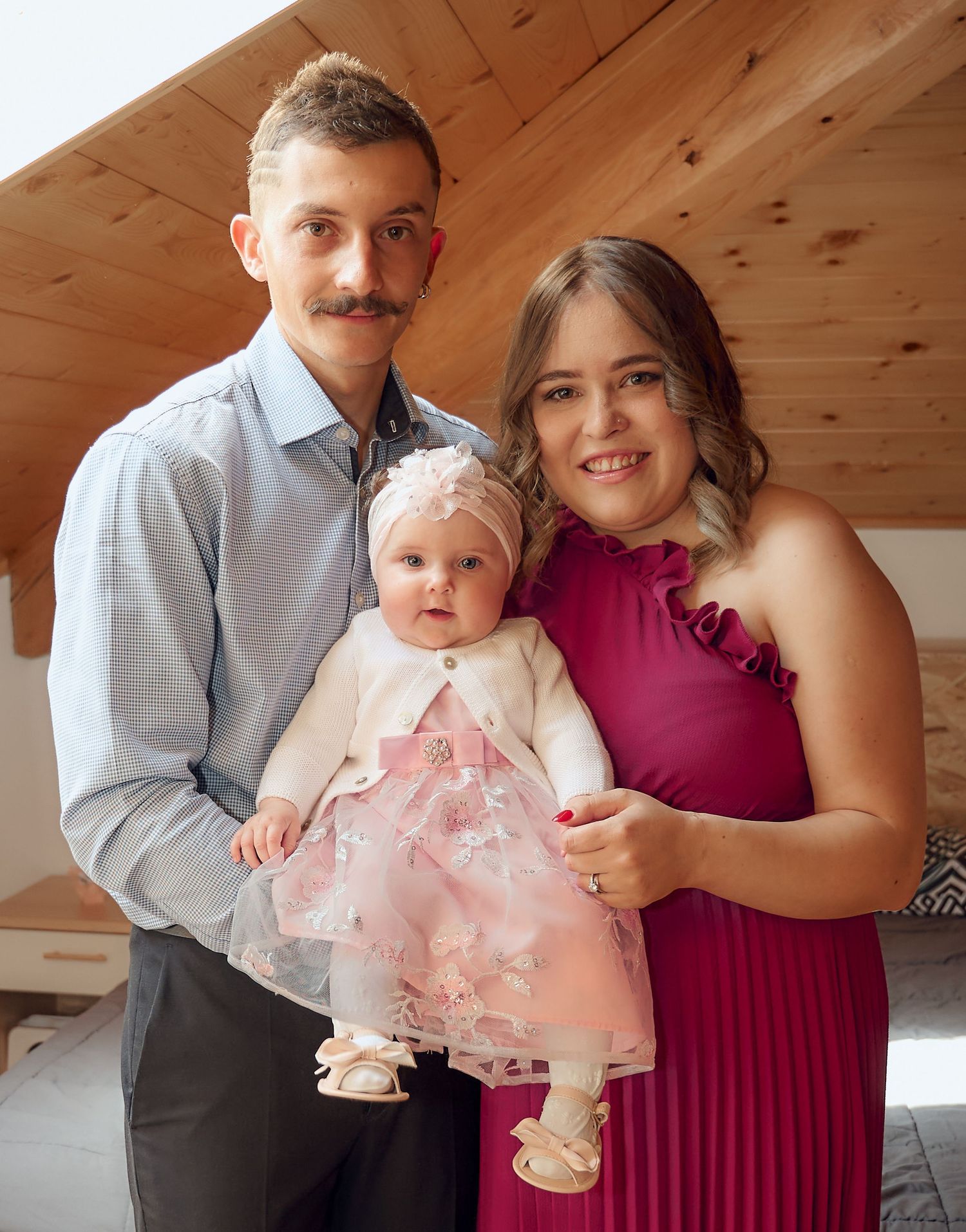 Happy family portrait with father, mother in magenta dress, and baby girl in pink baptism dress.