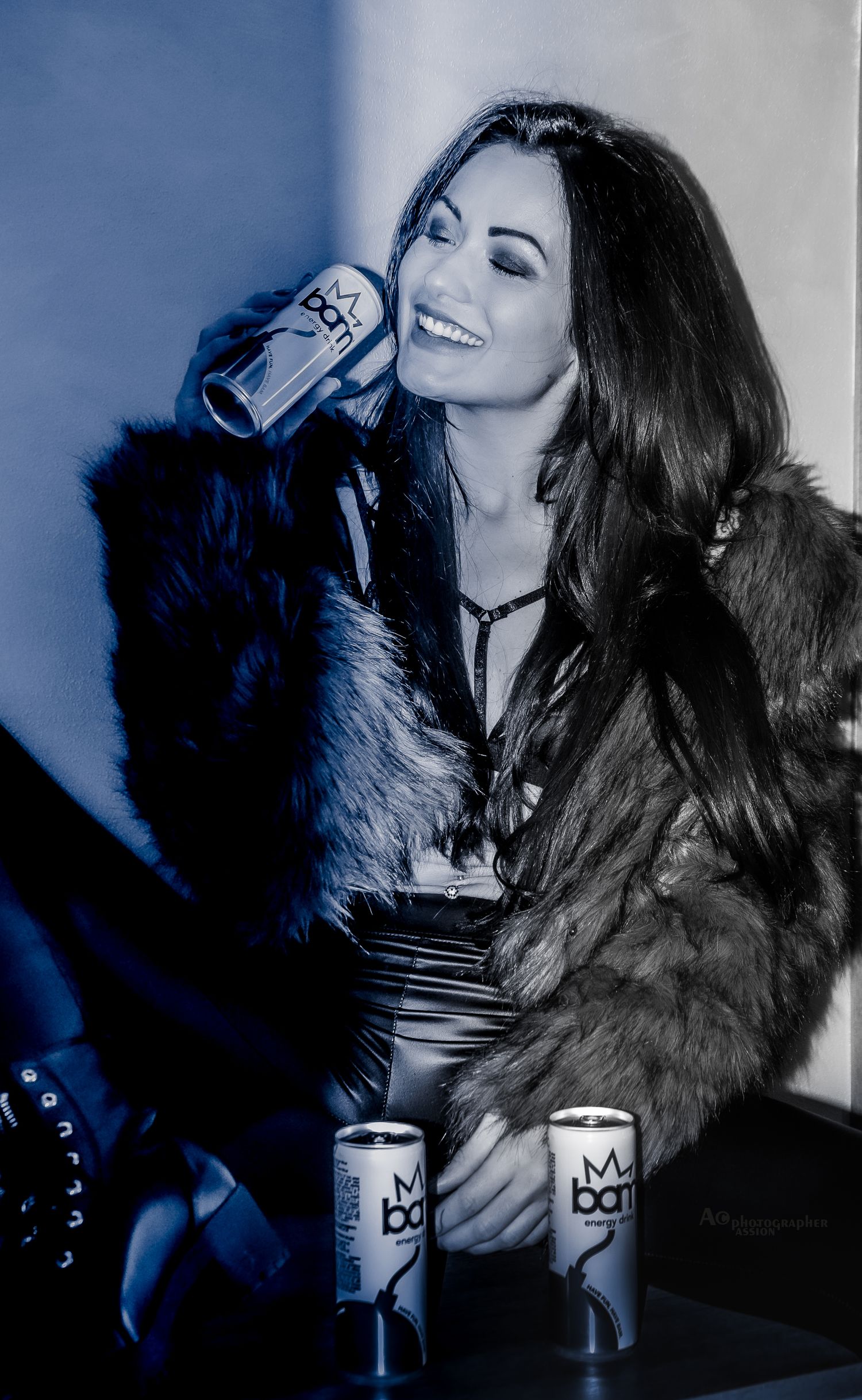 Woman in fur coat laughing while holding Bang energy drink in blue-toned black and white photo.