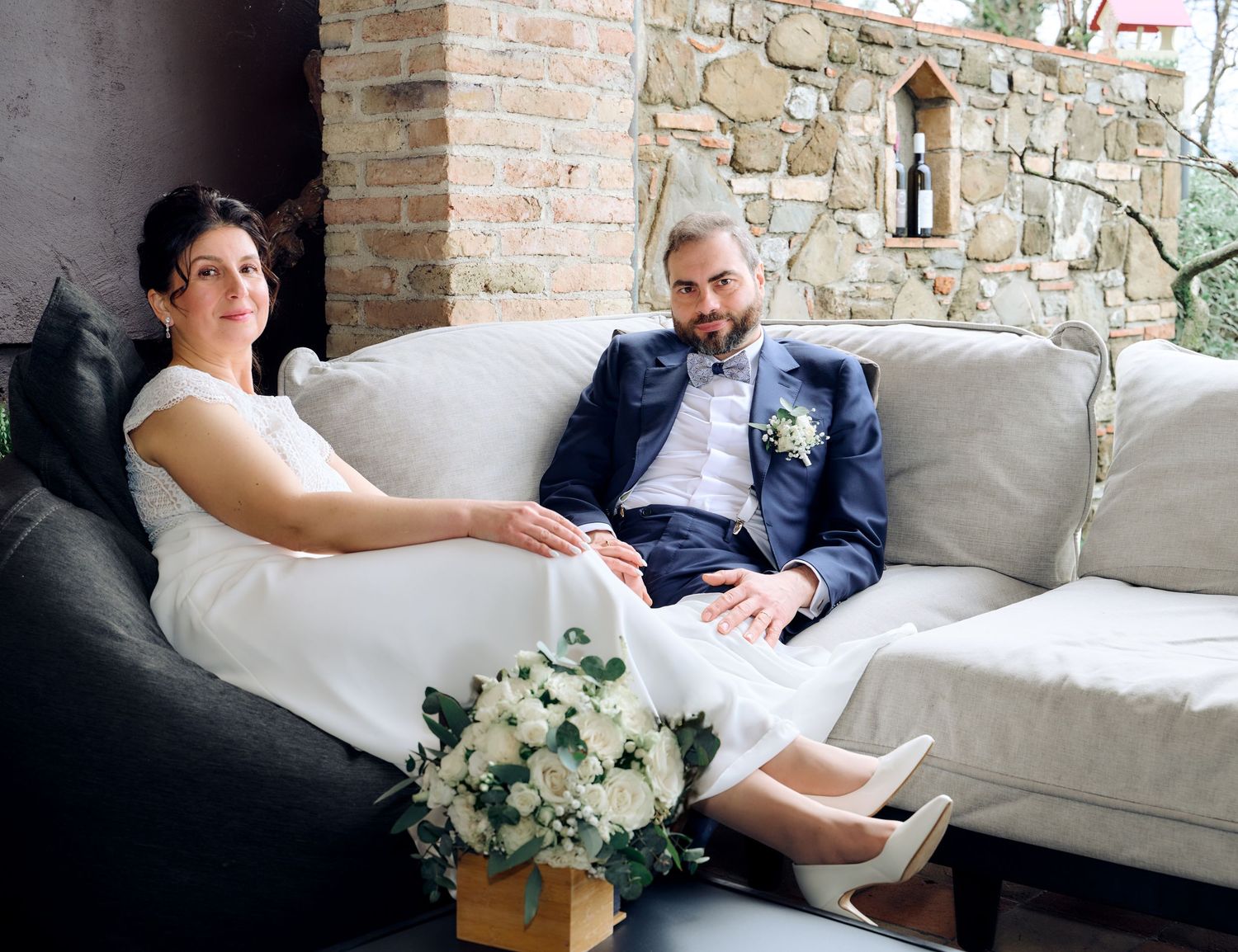 Bride and groom relax together on white sofa with floral bouquet in rustic stone venue setting.
