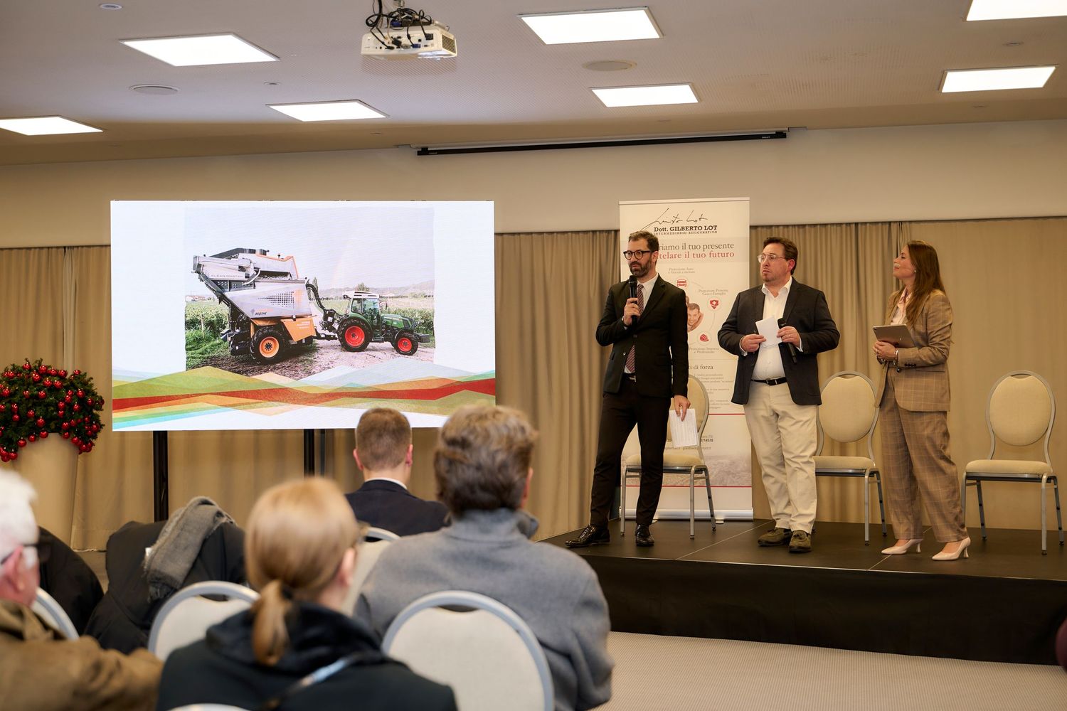 Two presenters speaking at a conference with an agricultural machinery slide projected behind them.