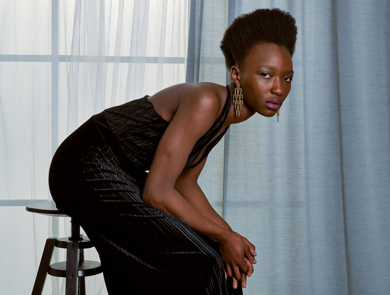 An elegant Black woman in a black sequined dress poses confidently on a stool indoors.