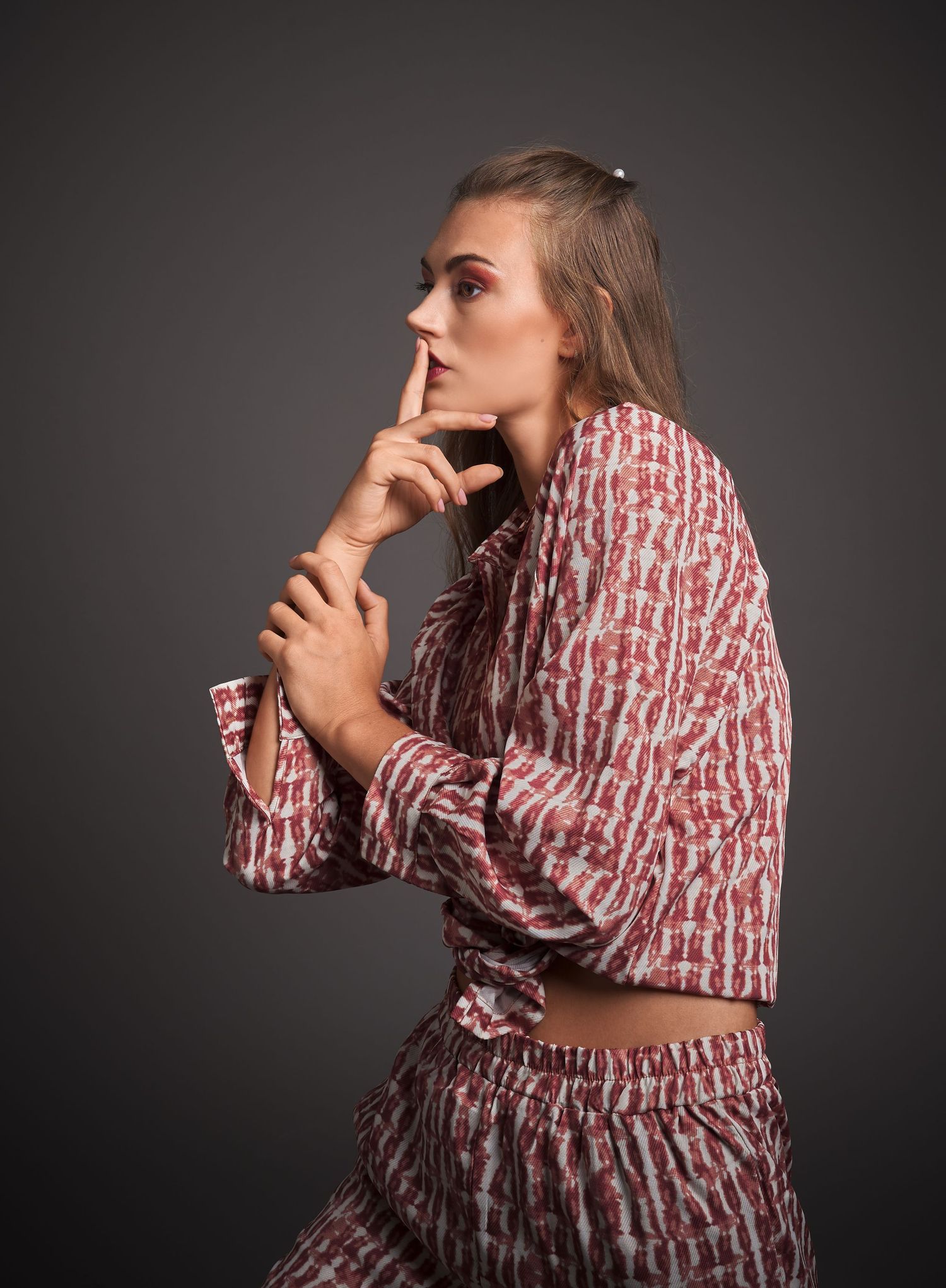 Elegant woman in patterned red outfit posing thoughtfully with finger on lips against gray background.