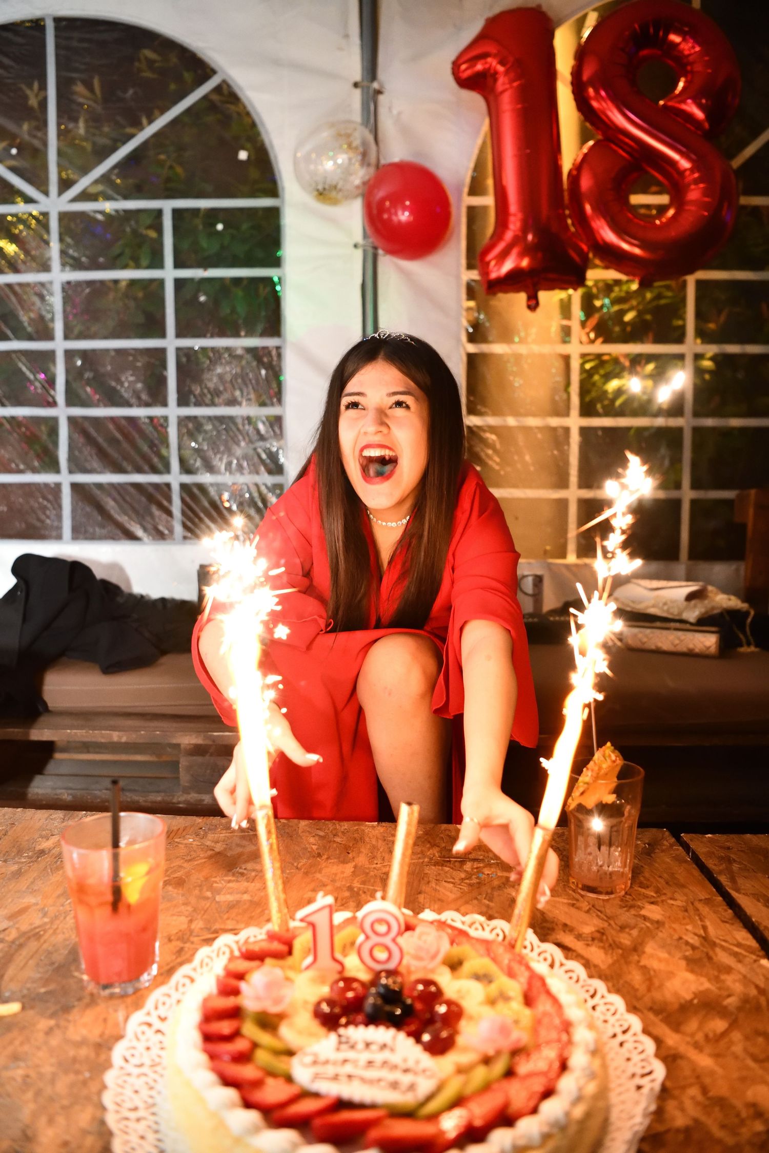 Excited young woman in red dress with sparklers beside her 18th birthday fruit cake.