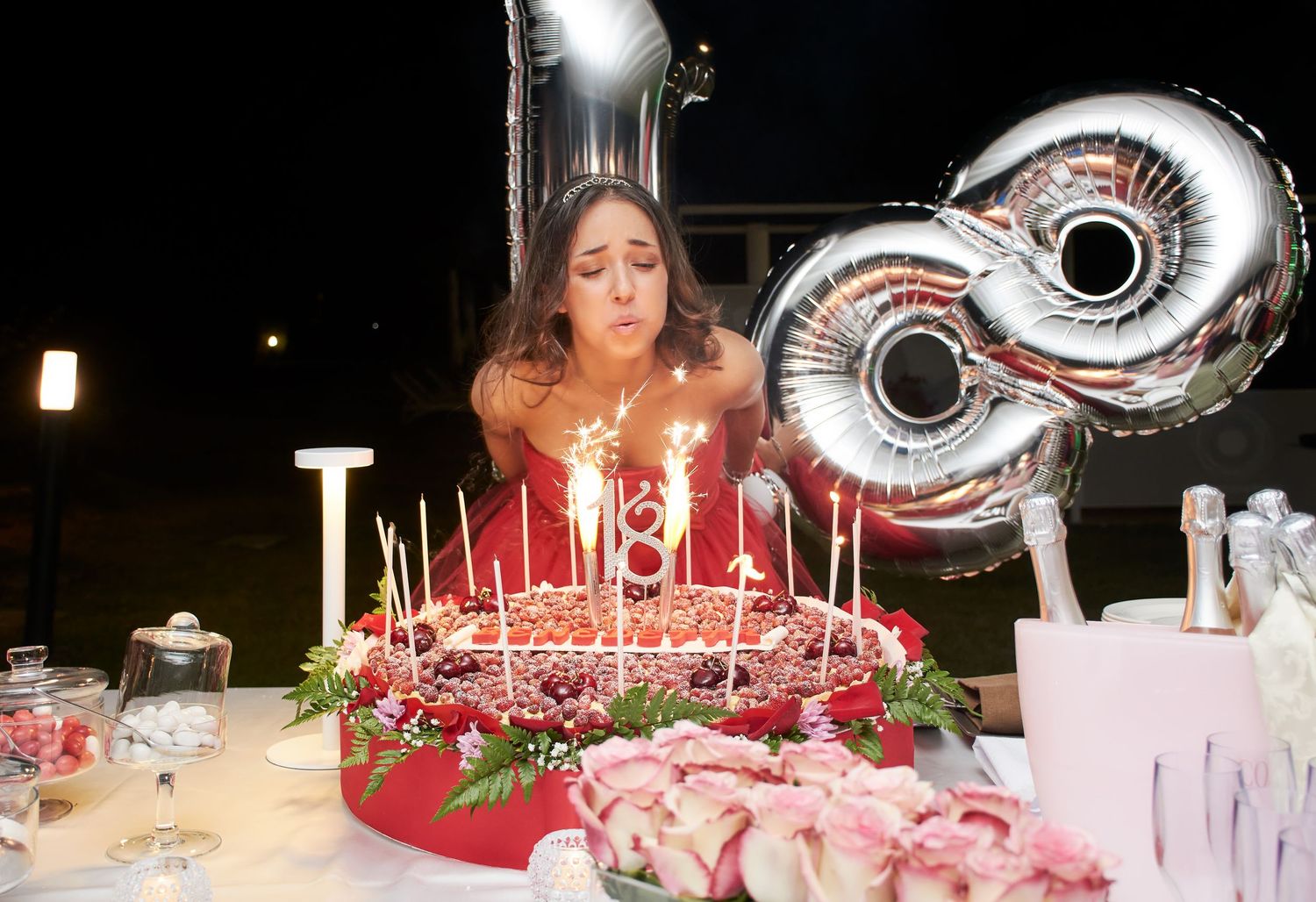 Young woman blowing out candles on an 18th birthday cake with sparklers at night.
