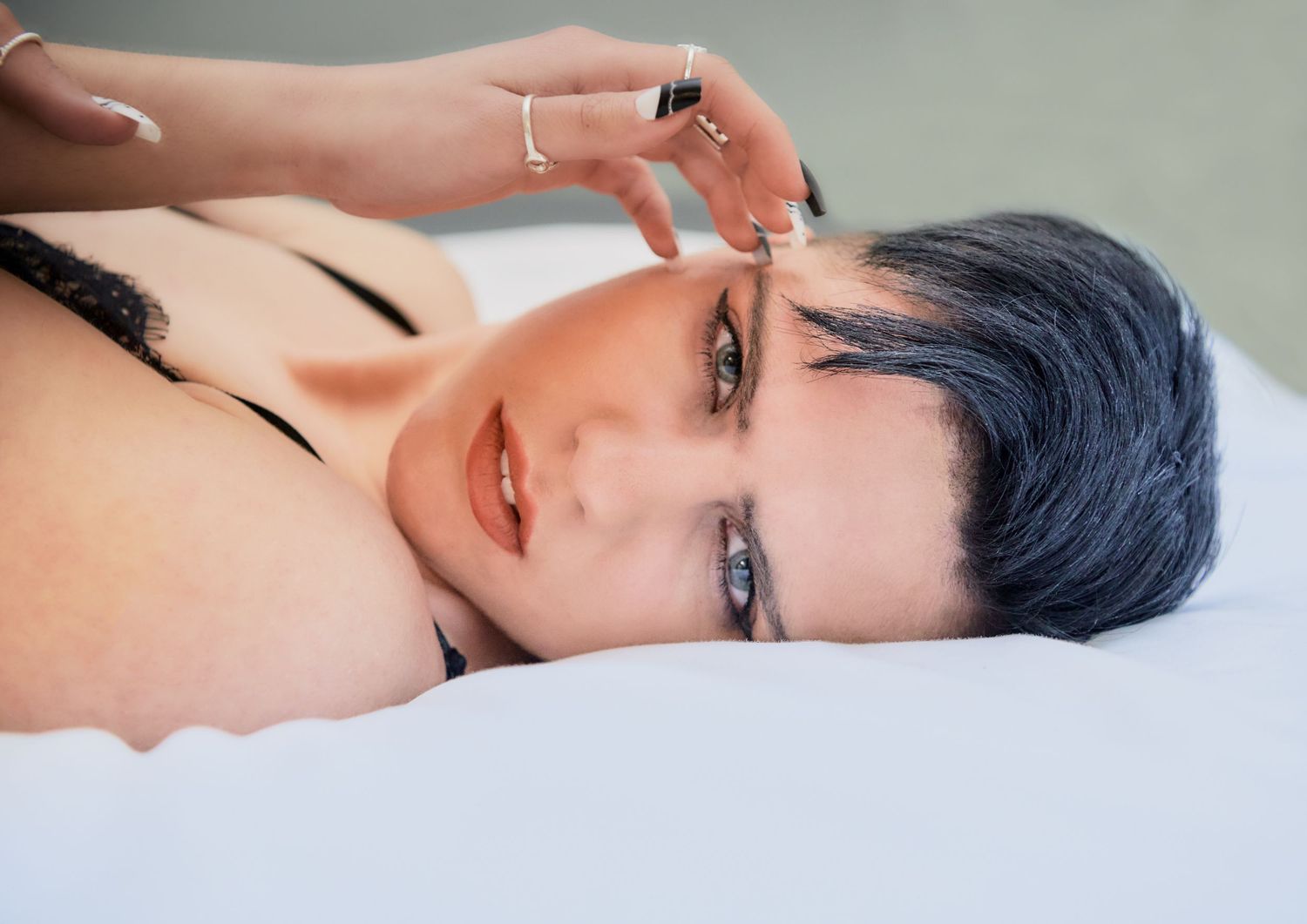 Short blue-haired woman lying on white surface wearing black bralette with rings.