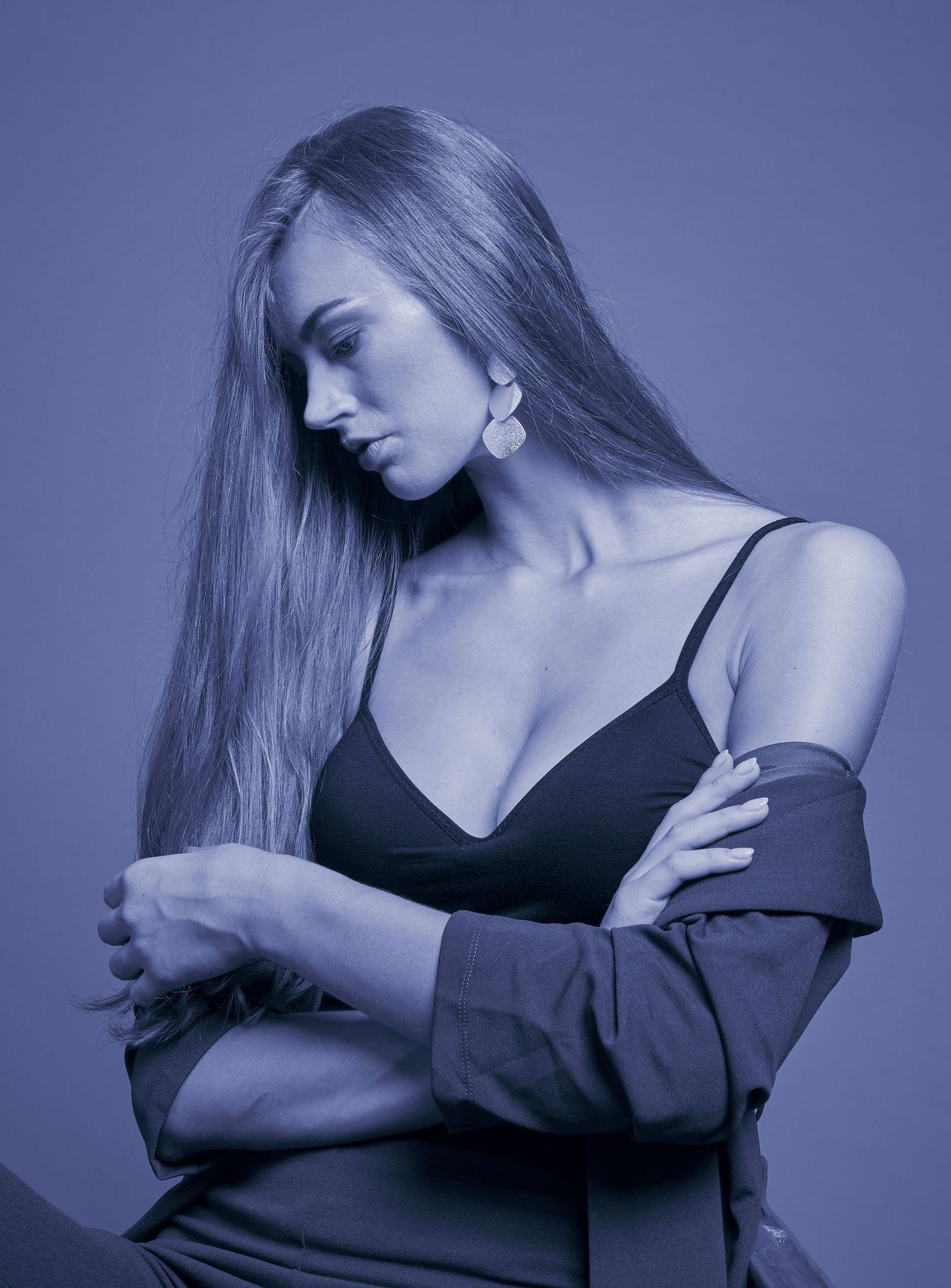 Blue-toned studio portrait of blonde woman in black bralette with arms crossed looking downward.