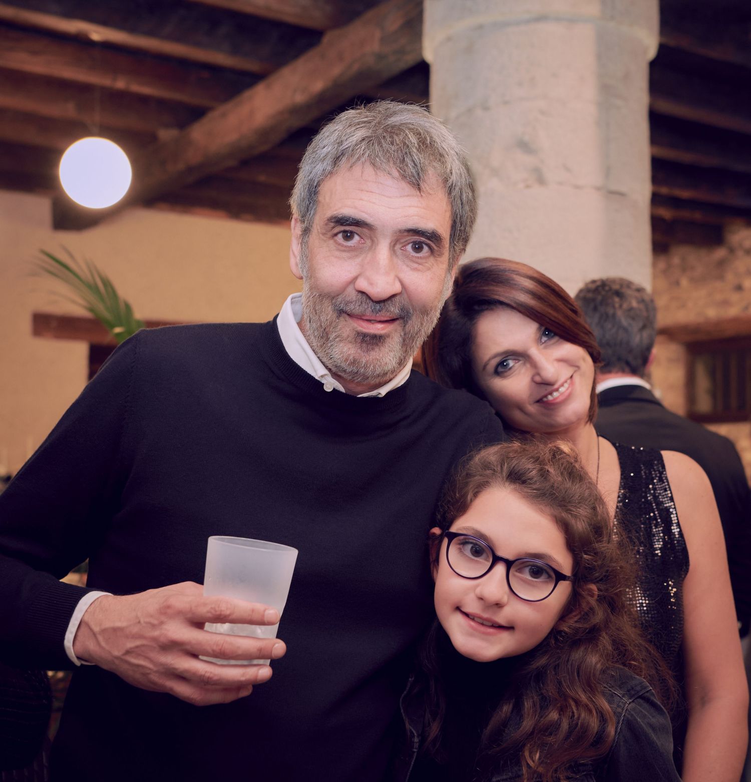 A man holding a drink poses with a woman and young girl wearing glasses at an indoor evening event.