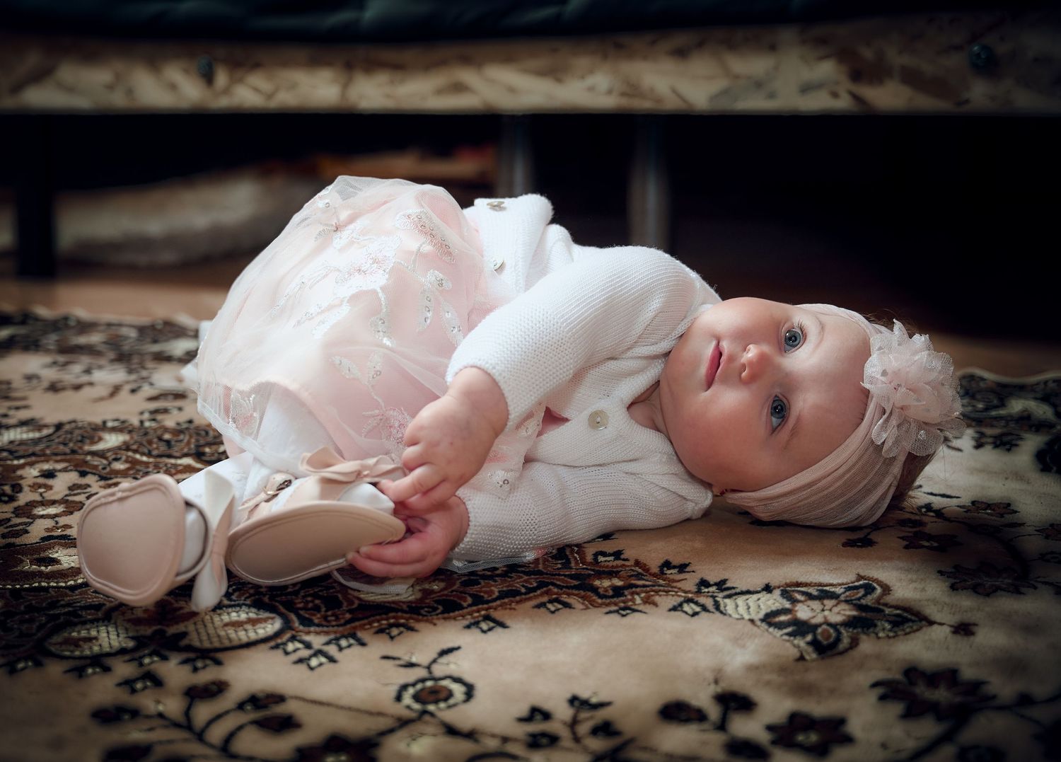 Beautiful baby girl in white cardigan and pink tutu lying on vintage patterned carpet.