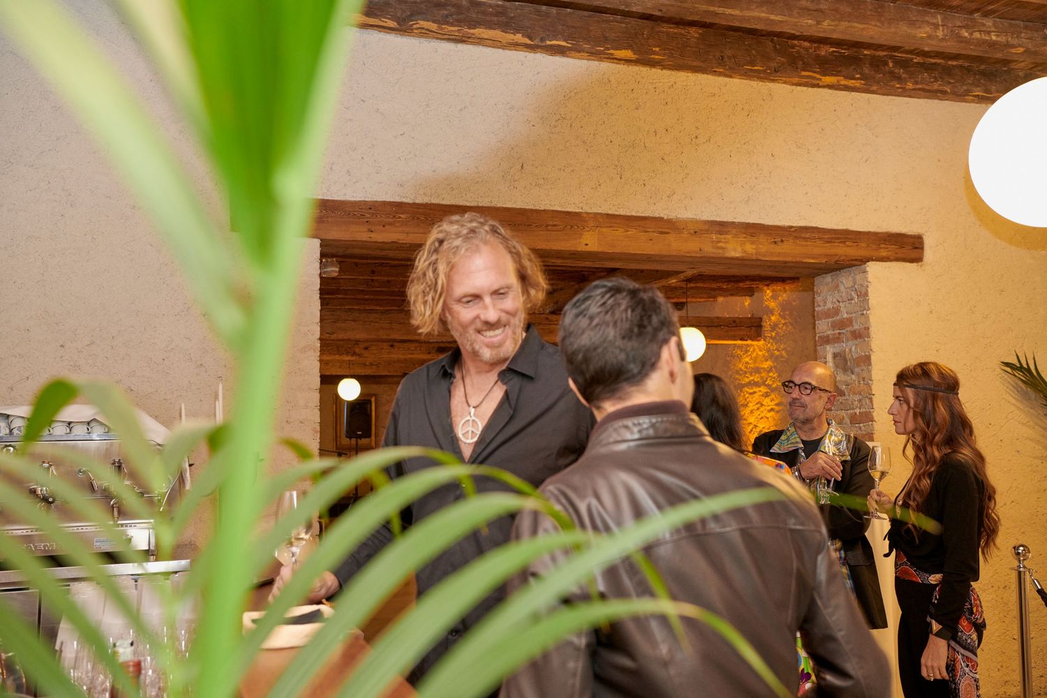 A curly-haired man laughs while conversing with guests near tropical plants at an indoor party.