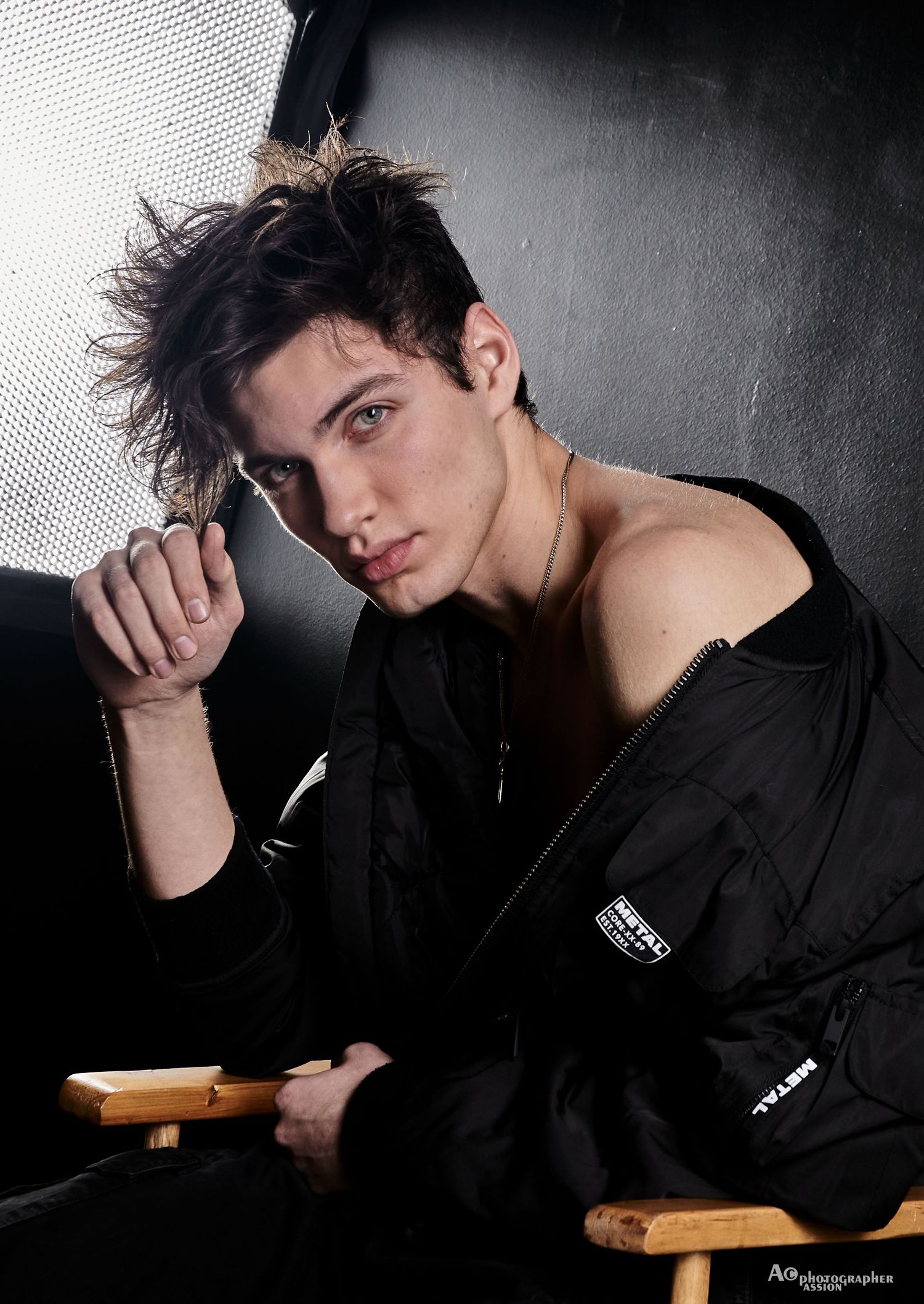Young male model with tousled dark hair wearing black bomber jacket seated in director chair.