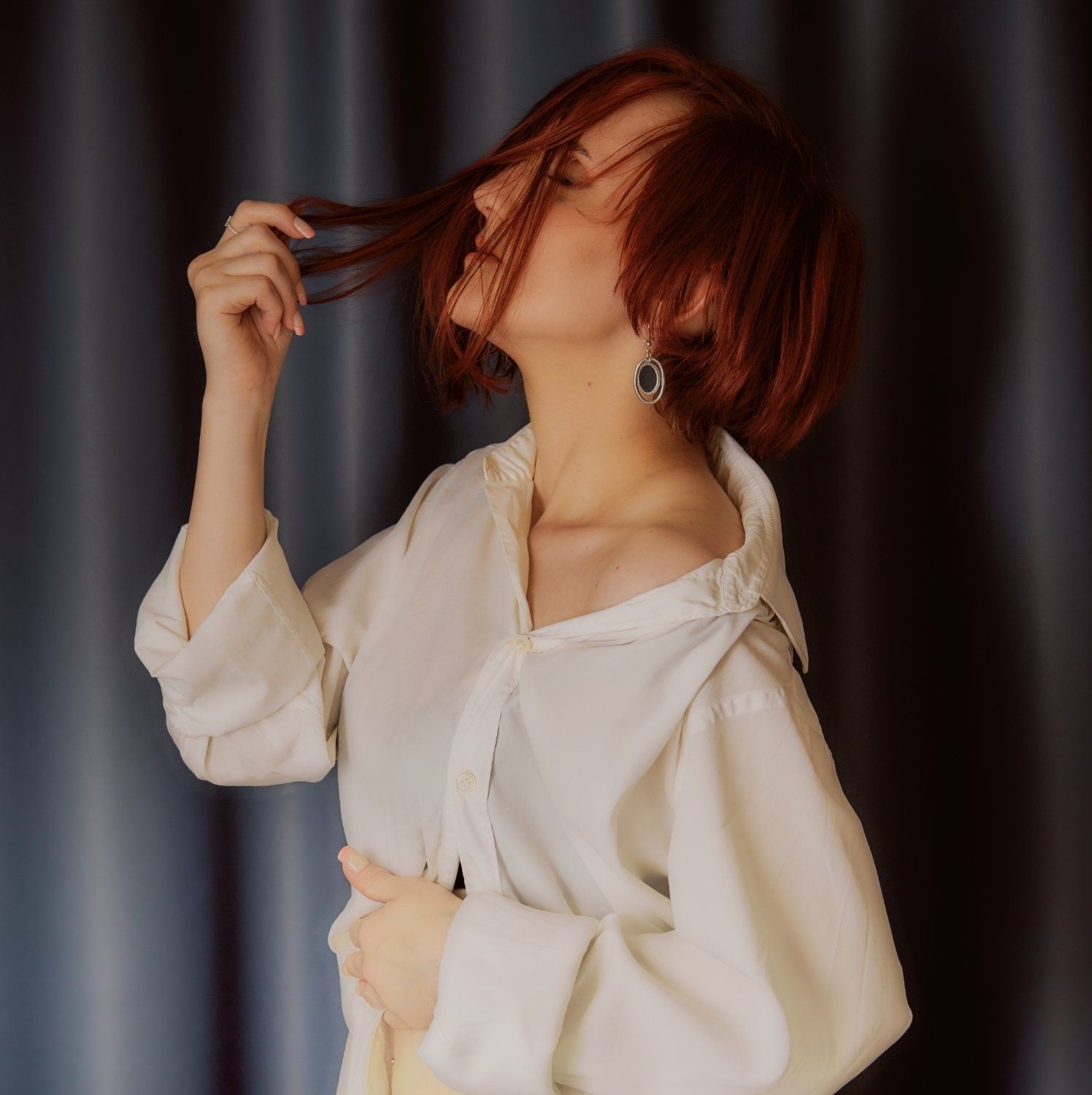Redhead woman in oversized white shirt poses with eyes closed, touching her hair against curtain backdrop.