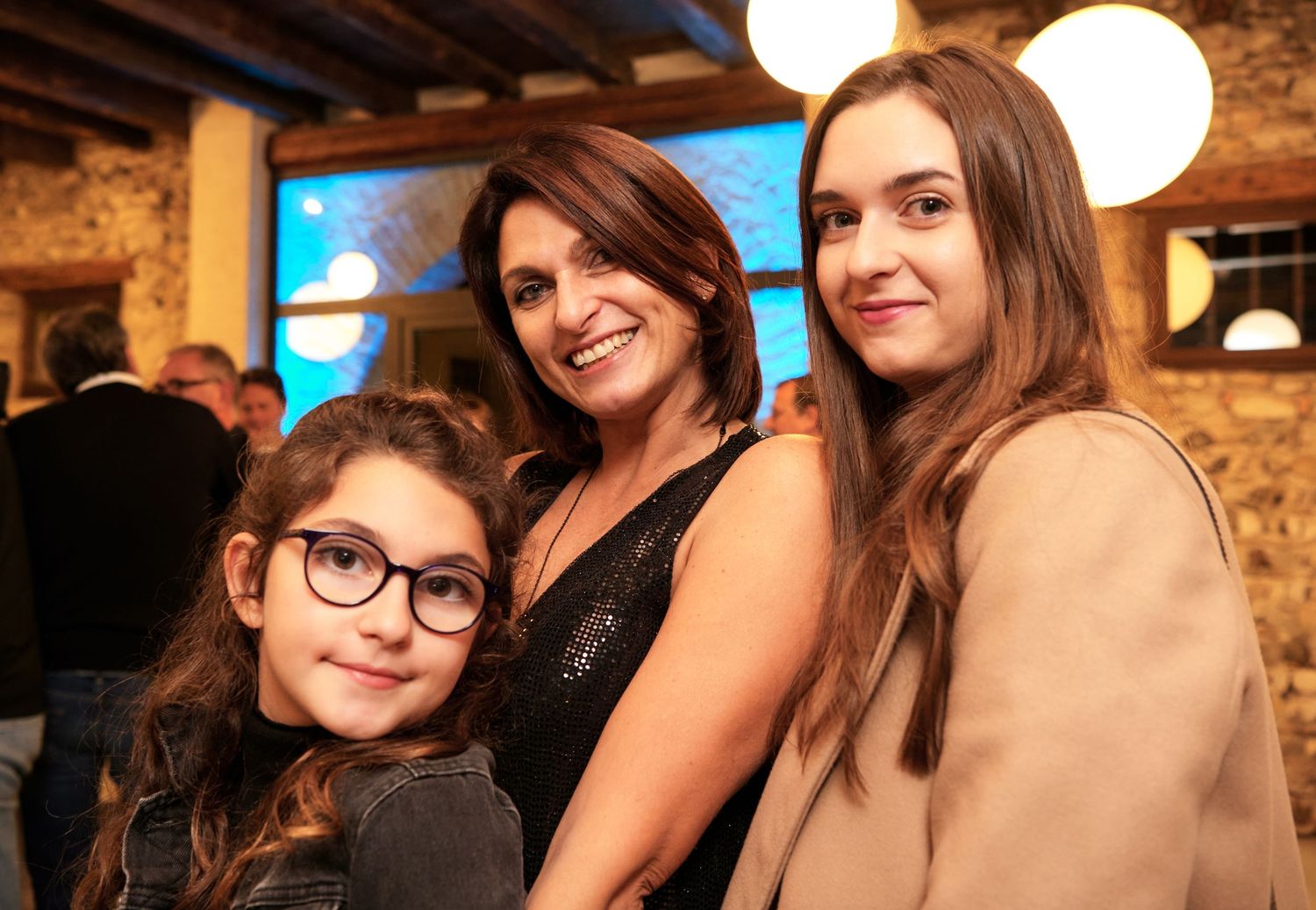 A woman poses warmly with two girls, one wearing glasses, at an indoor evening gathering.