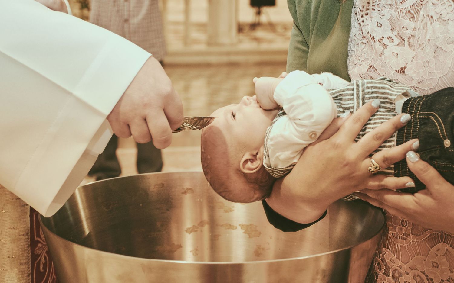 Catholic priest performing infant baptism ceremony, pouring holy water over baby's head at baptismal font.