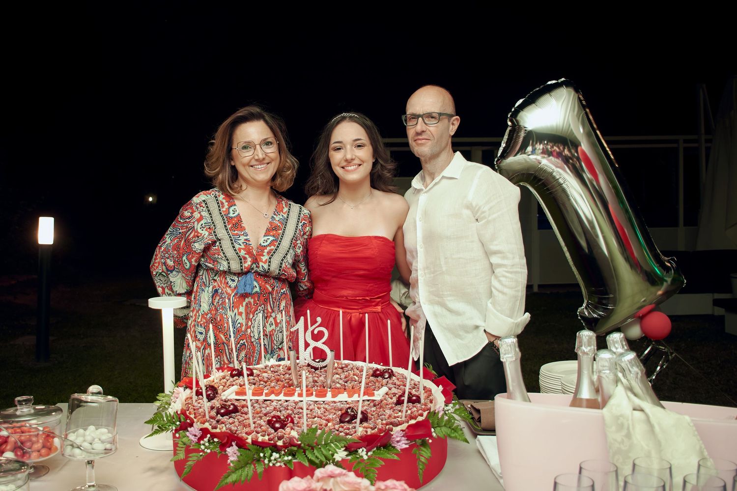 Family of three posing with an 18th birthday cake and silver balloon decorations at night.